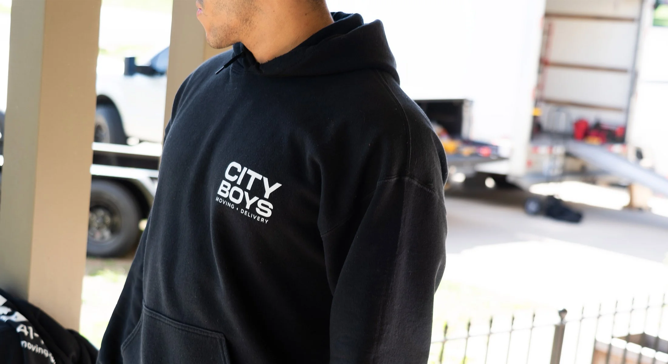A person is wearing a black hoodie with a logo that reads 'CITY BOYS MOVING, DELIVERY' on the front. They are outdoors near a warehouse or moving truck, with some equipment and cars visible in the background.