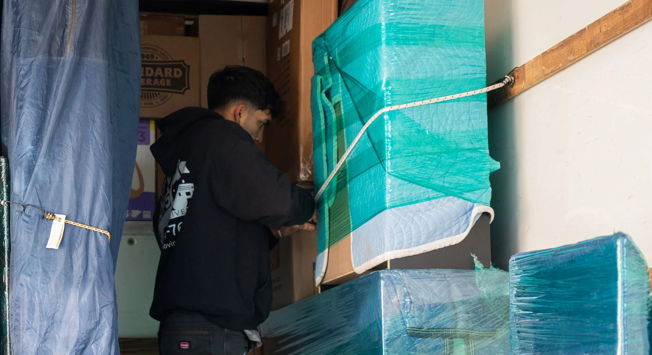 A person with dark hair and a black hoodie is working in a storage or warehouse area, surrounded by large wrapped objects and boxes.