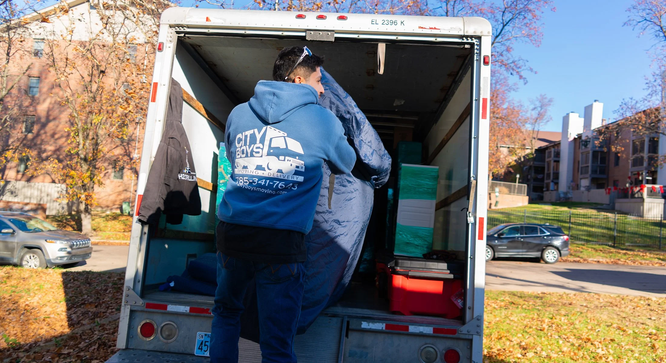 A man loading moving boxes into the back of a moving truck on a sunny day in a residential neighborhood with trees and multi-story apartments in the background.