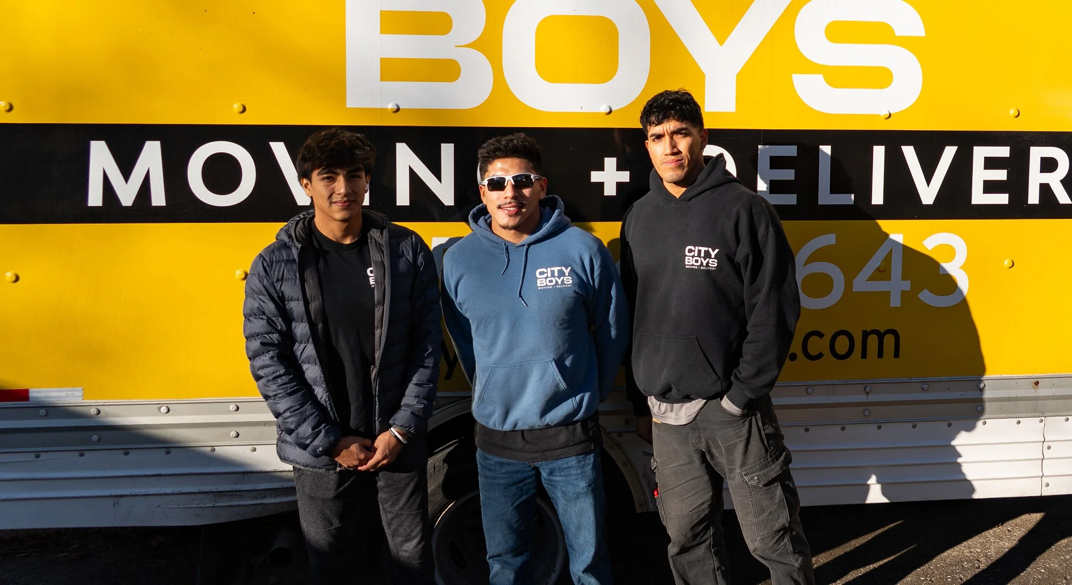 Three young men standing in front of a yellow and black moving truck with the company name 'City Boys' and slogan 'Moving + Delivery' visible on the truck. They are wearing casual clothing and sunglasses, posing for the photo with a sunny background.