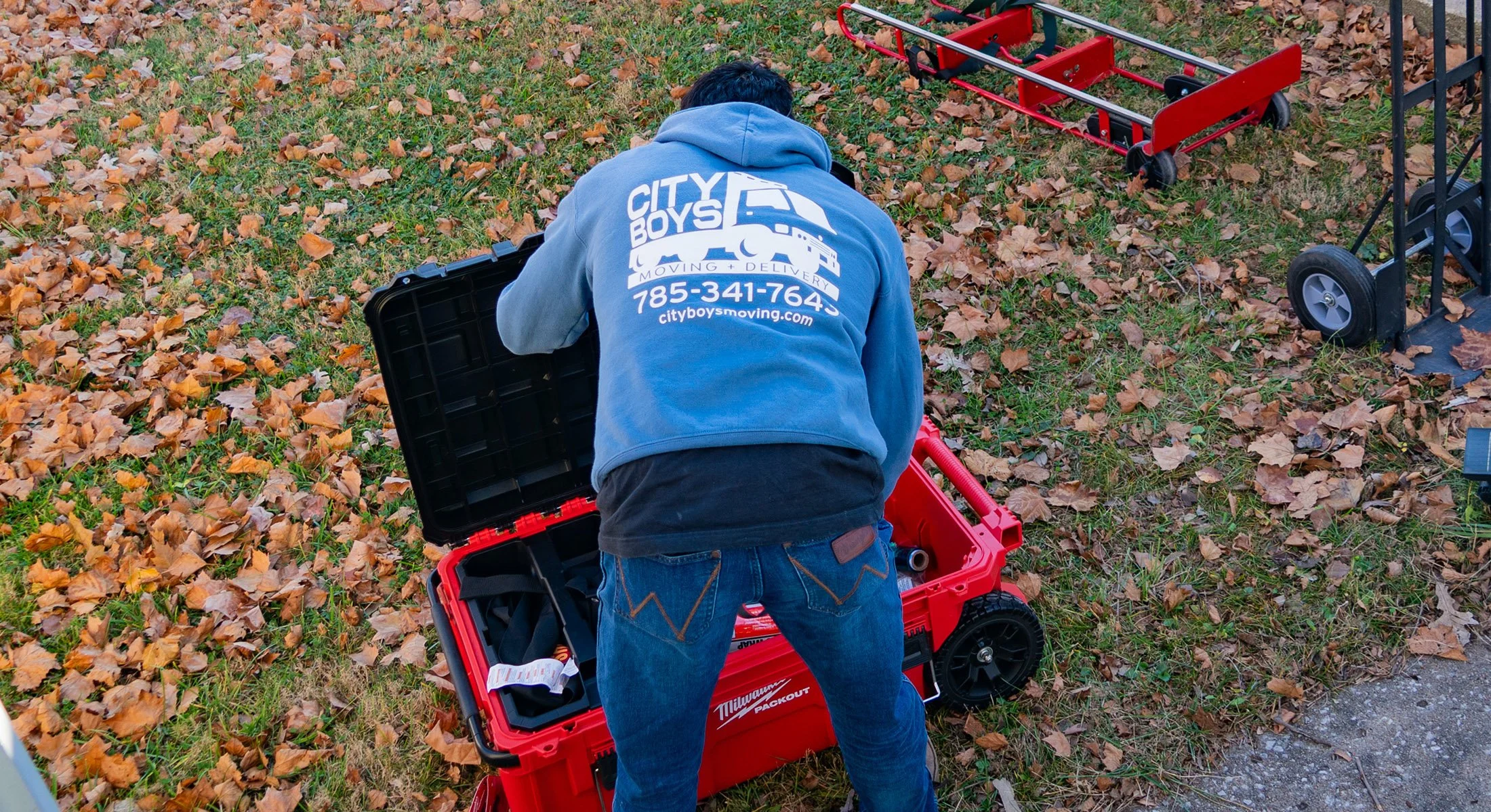 Person in a blue hoodie with 'CITY BOYS MOVING' on the back, kneeling next to a red toolbox and a wagon, outdoors on a grassy area covered with fallen leaves, surrounded by tools and equipment.