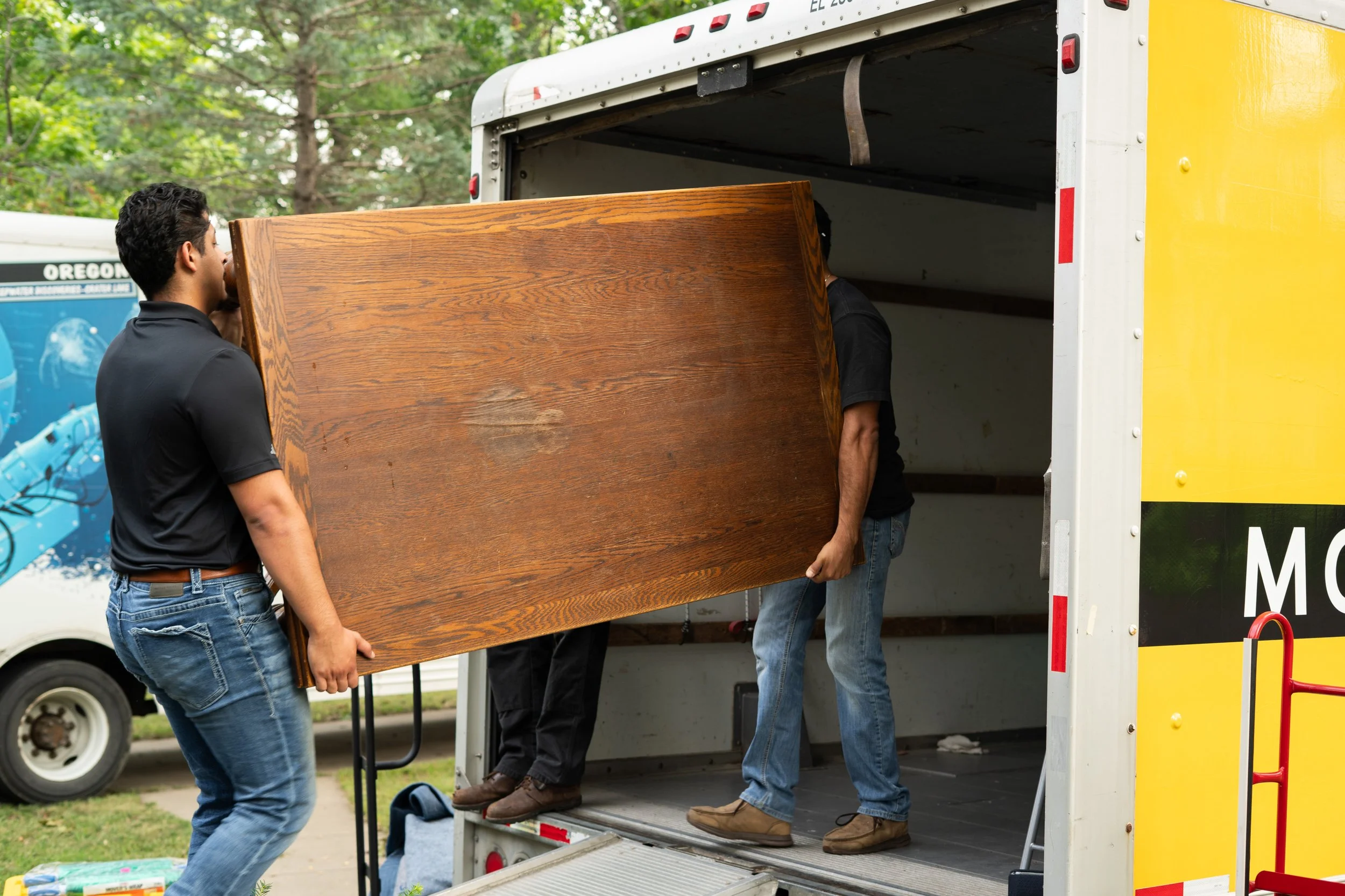People loading a wooden furniture piece into a yellow moving truck with the back open, outdoors surrounded by trees.