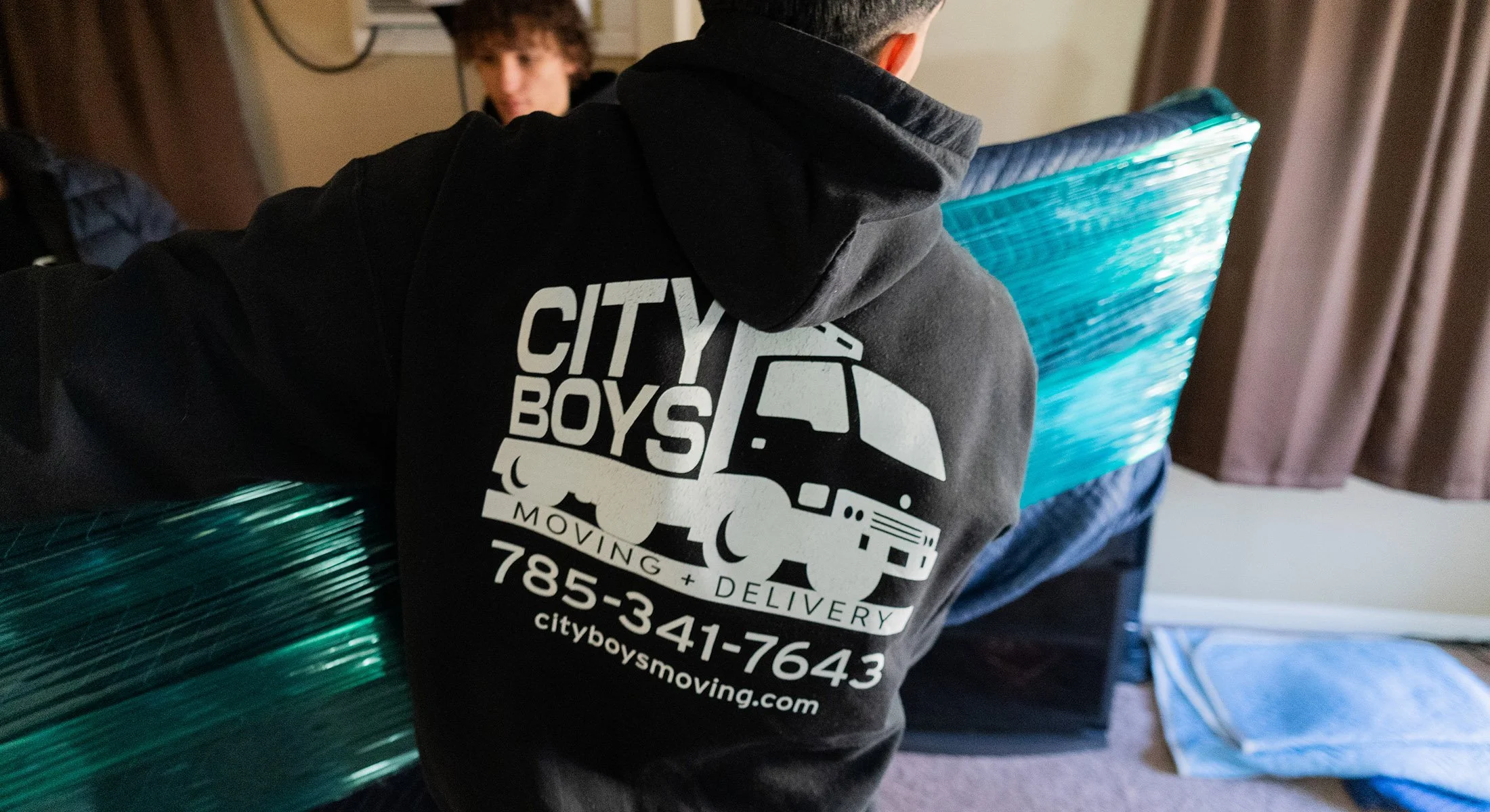 Person wearing a black hoodie with white text and a logo for City Boys Moving, carrying a large bundle of green metal pipes in an indoor setting.