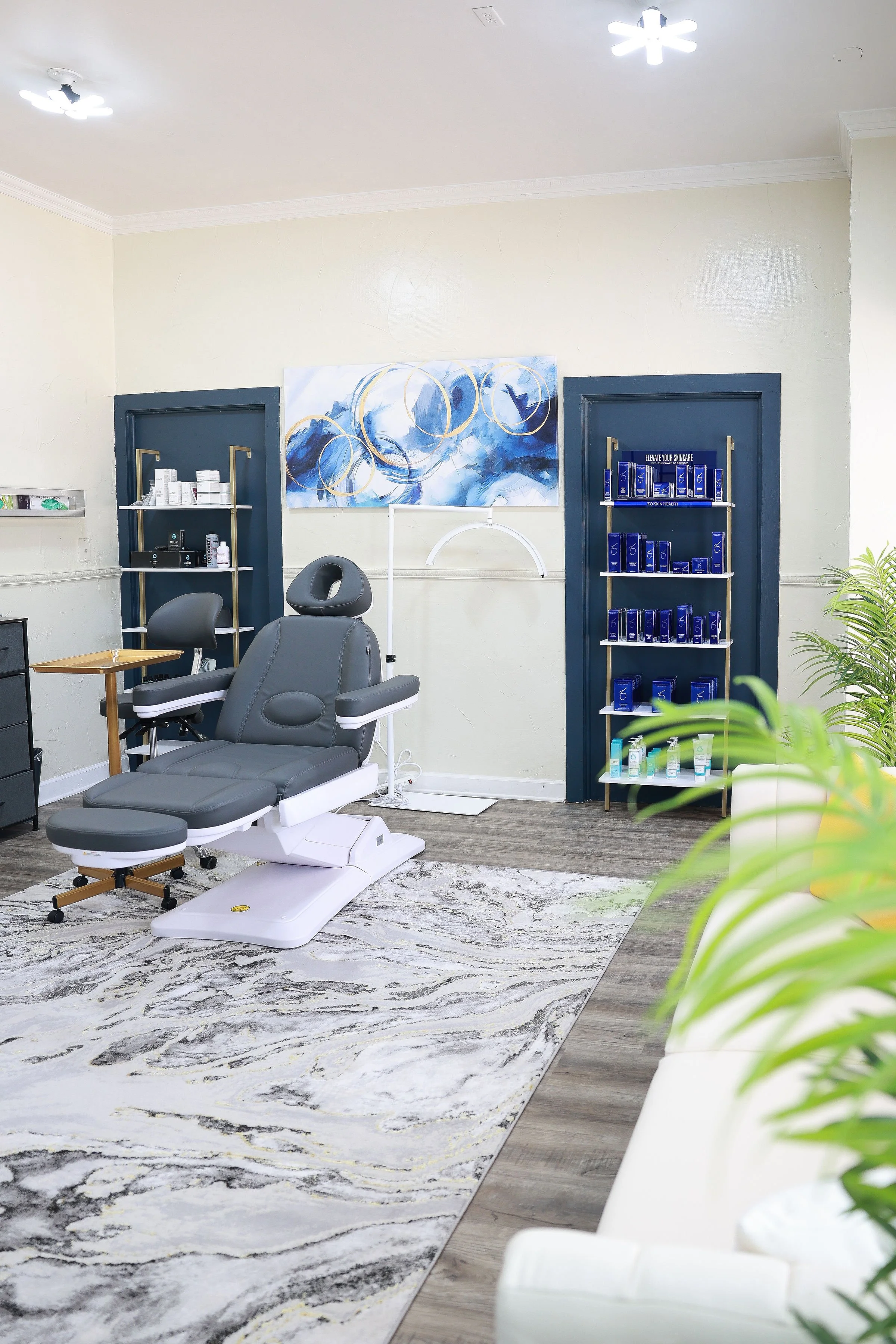 A medical or beauty treatment room with a gray reclining chair, navy blue shelves stocked with skincare or medical products, a modern abstract painting, and a patterned rug on a wooden floor.
