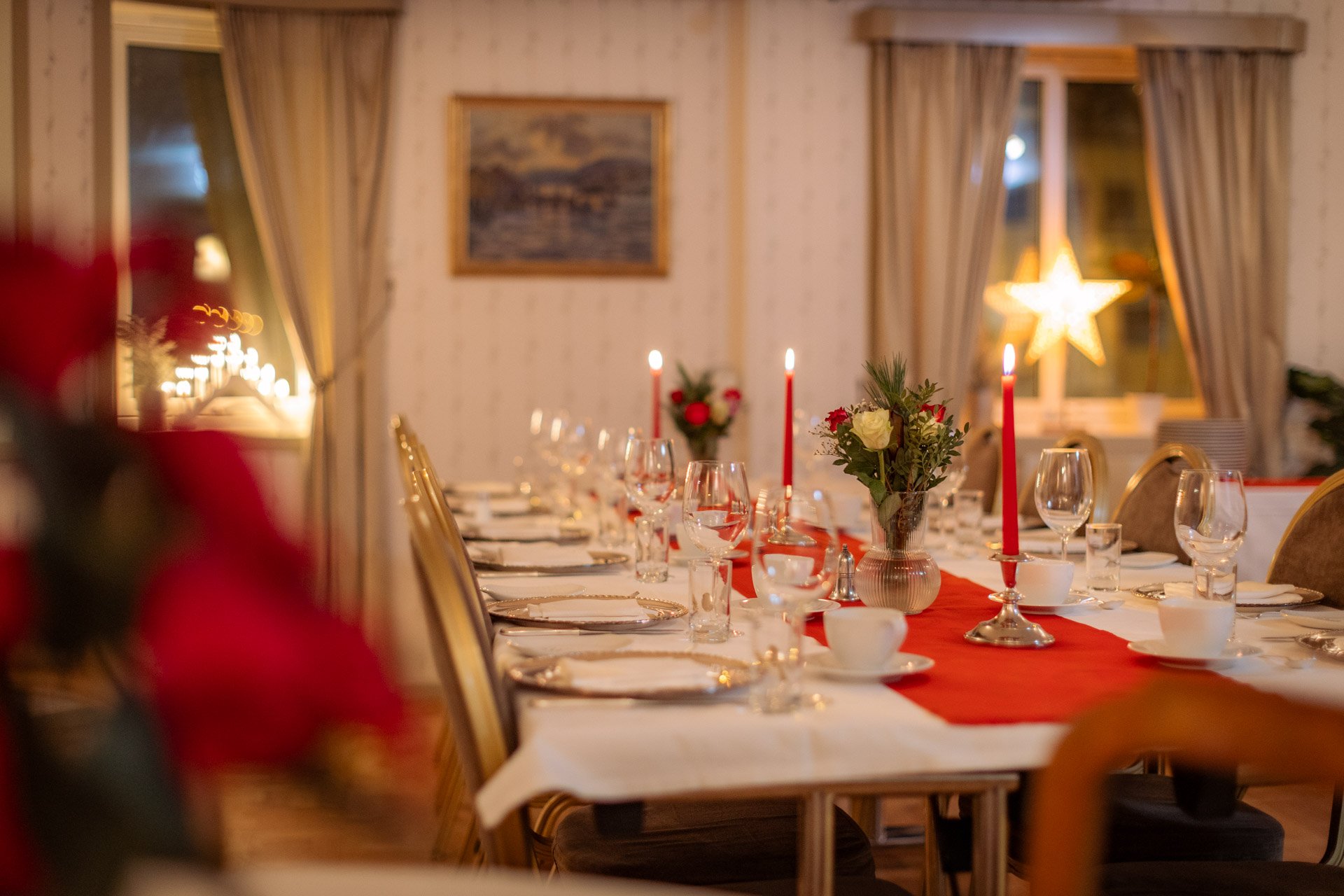 Festbord med kandelabra, blomsterdekorasjoner, glass, tallerkener, og bestikk i et pyntet rom med gardiner og et stjerneformet lyselement i vinduet.