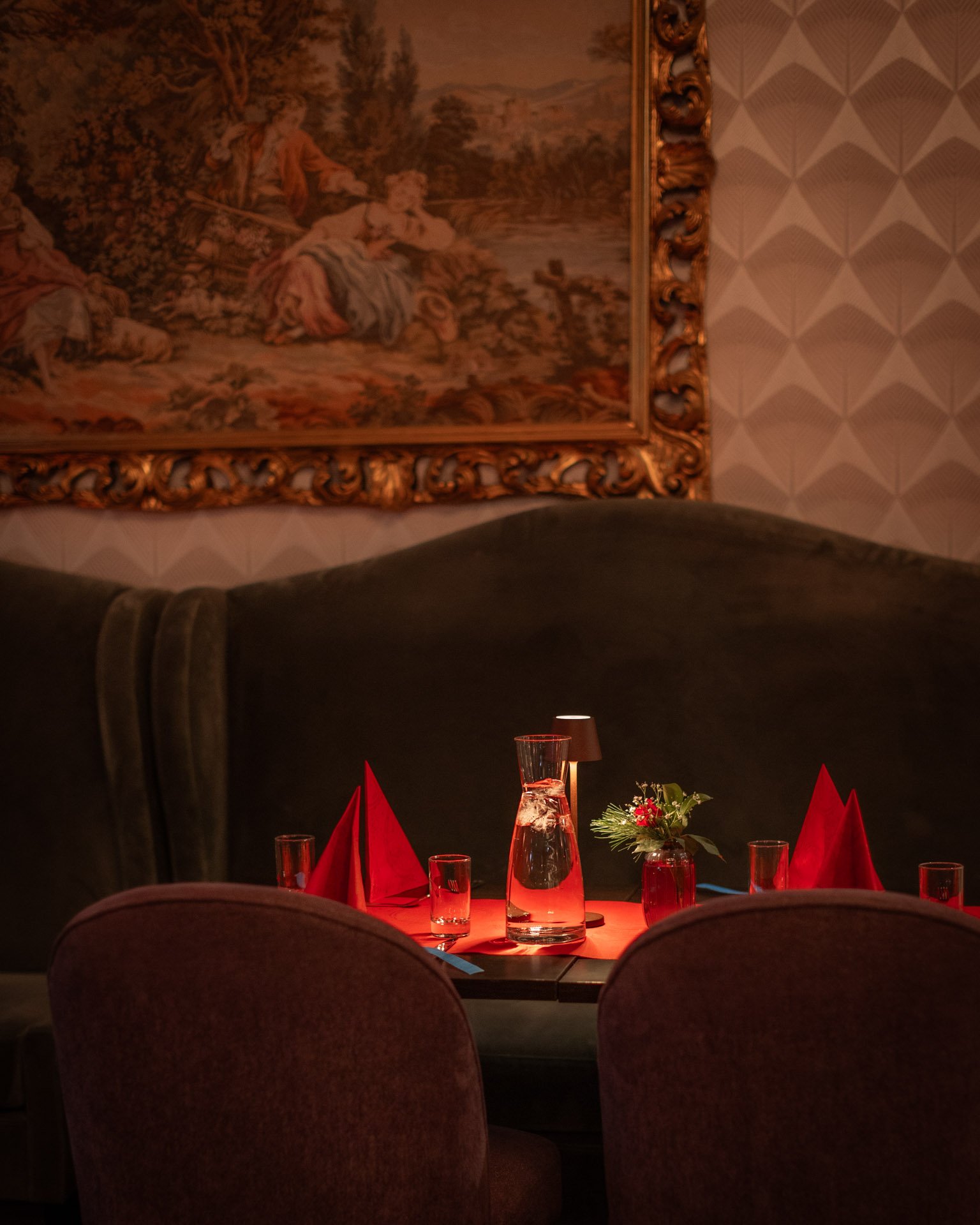 En restaurantscene med et bord dekket med røde servietter og vannglass, en glasskaraffel med vann, og en liten blomsterdekorasjon. Bakgrunnen viser et malt tapet og en rik, gullinnrammet kunstgalleri.