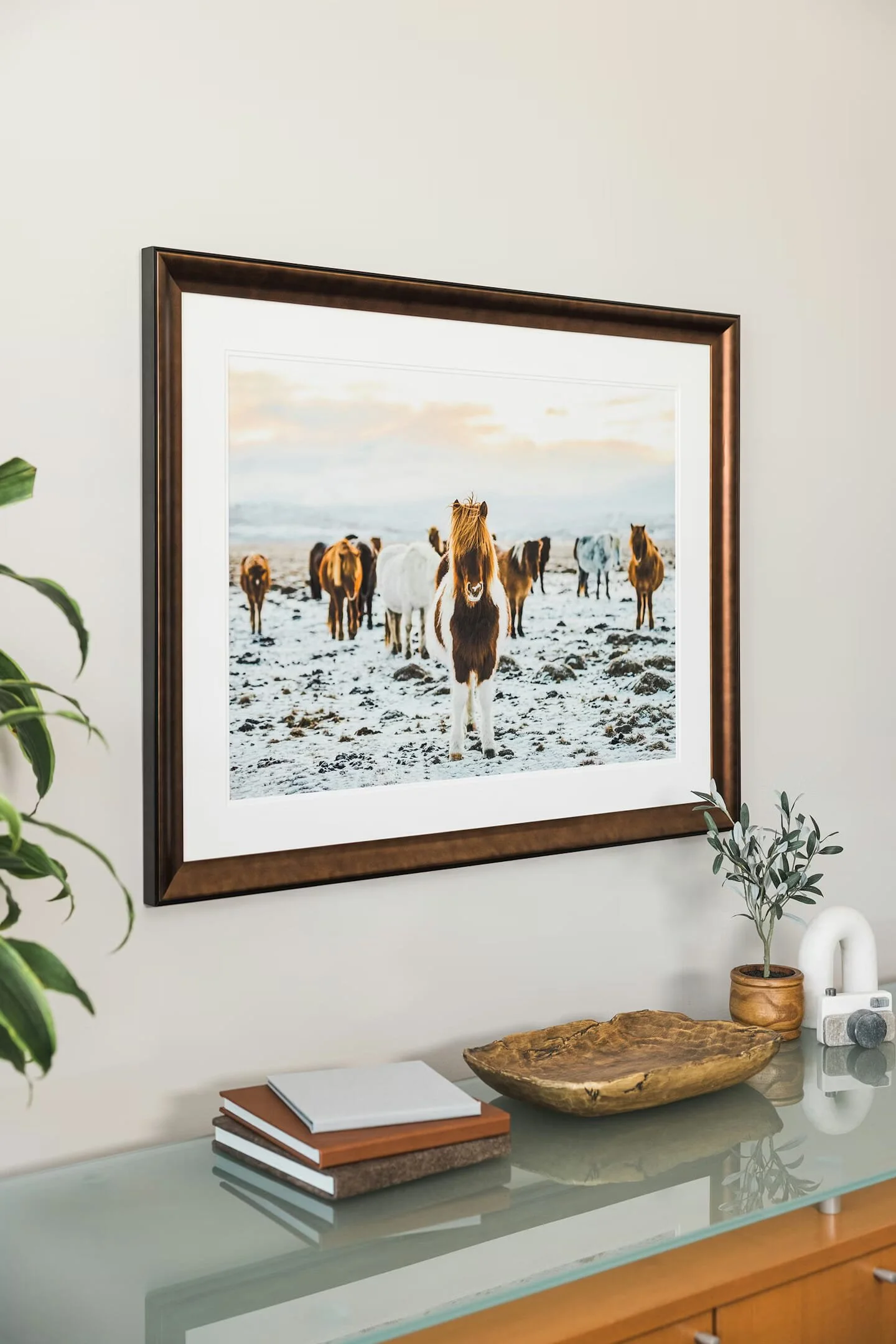 Framed photo of a group of horses standing on a sandy beach with a cloudy sky in the background, displayed on a wall above a glass-topped table with stacked notebooks, a wooden tray, a small potted plant, a white decorative object, and a camera.
