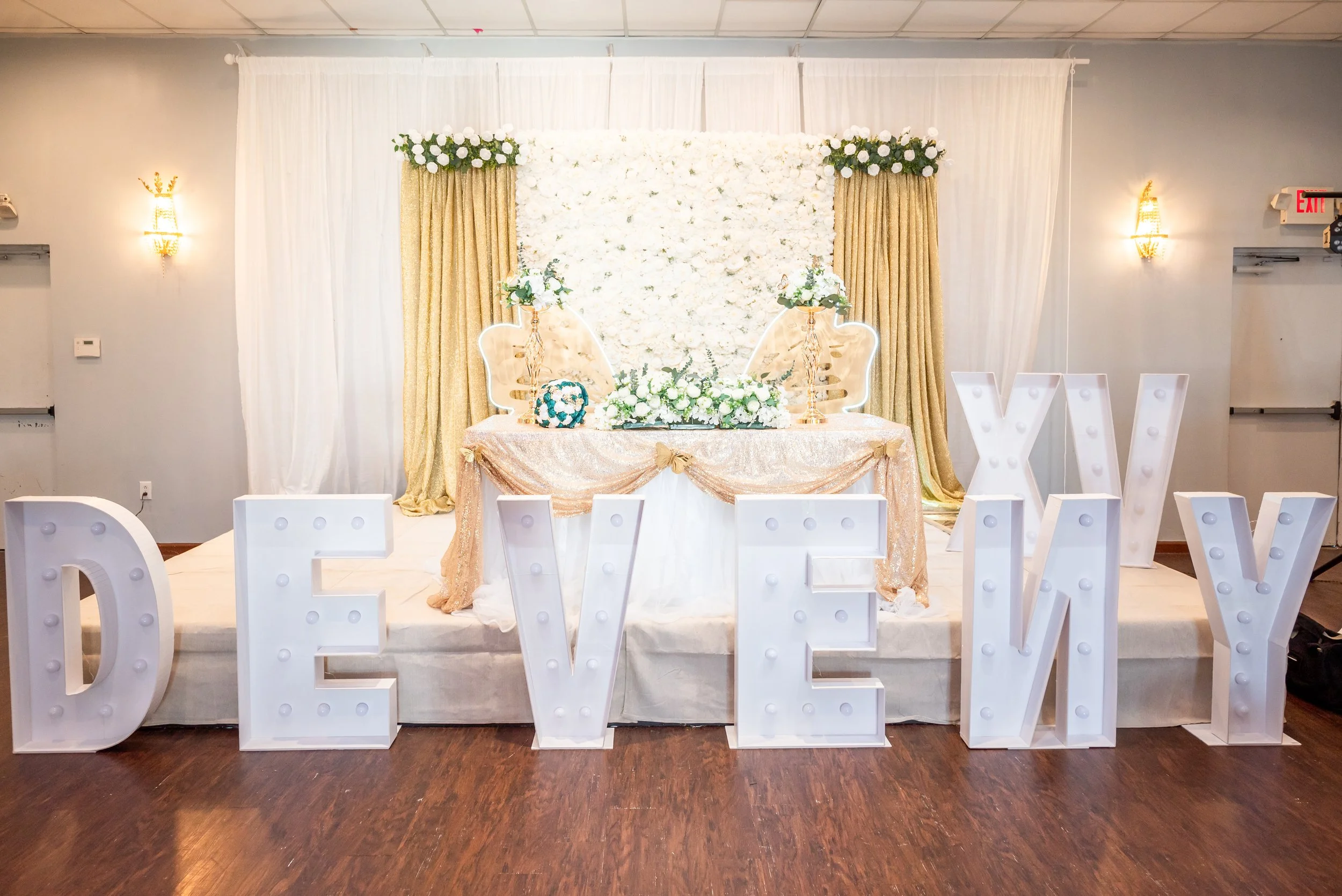 Decorated event stage with large illuminated letters spelling 'LOVEY' in front, a floral backdrop with white and gold drapes, and a table adorned with flowers and decorative chairs.
