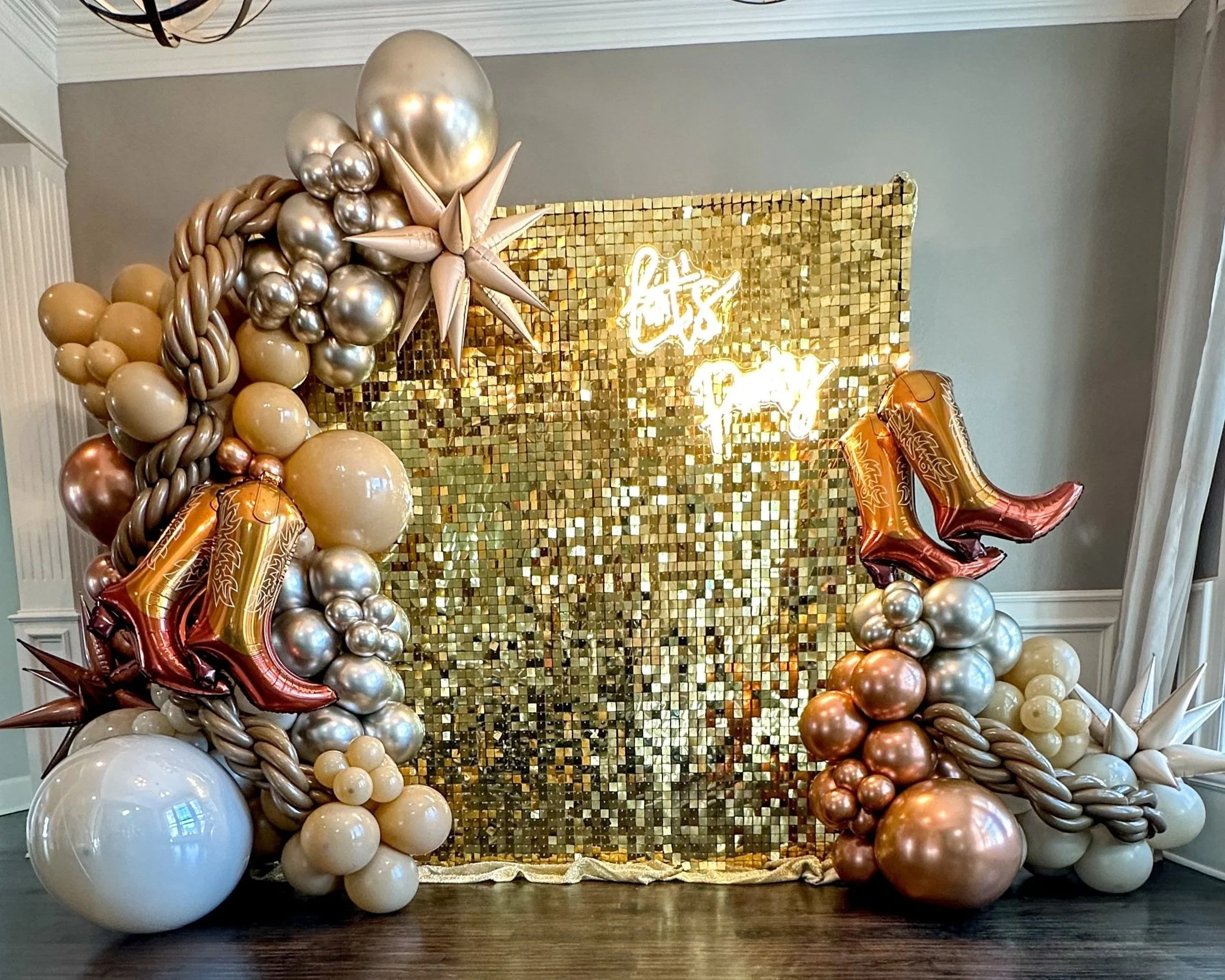 Decorative balloon arrangement with a gold mosaic backdrop and neon sign that says 'Let’s Party,' featuring large balloon cowboy boots and star accents.