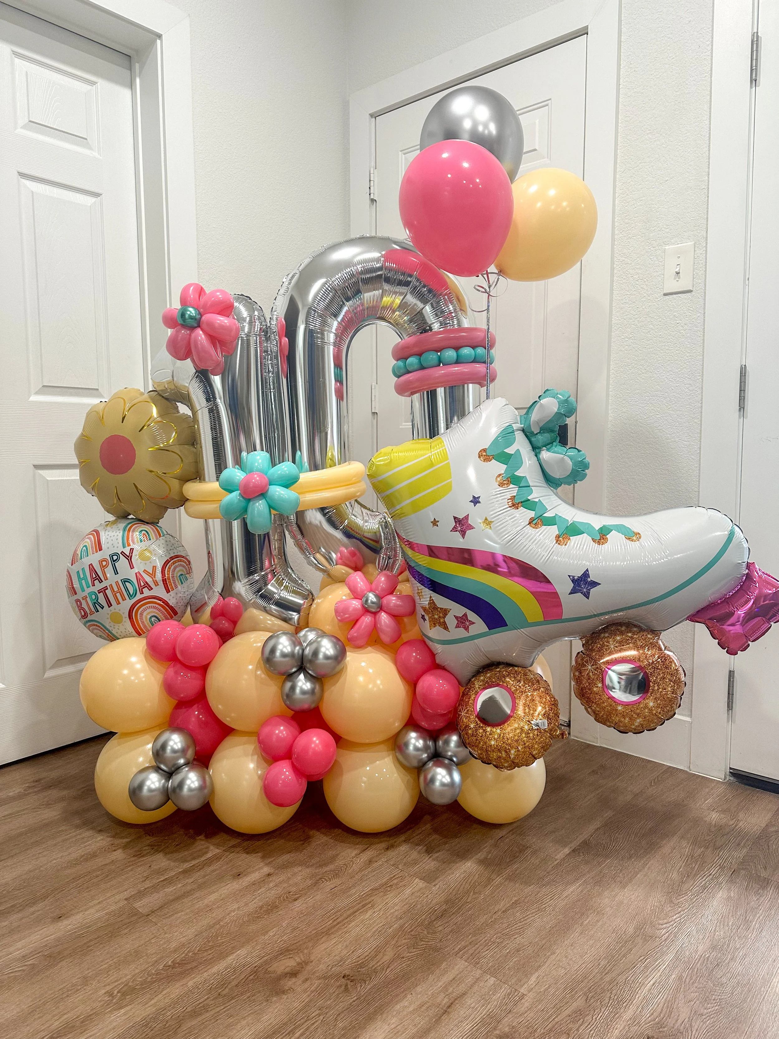 Colorful birthday balloon arrangement with large silver and pink balloons, a unicorn-shaped balloon, flower-shaped balloons, and a 'Happy Birthday' balloon on a wooden floor.