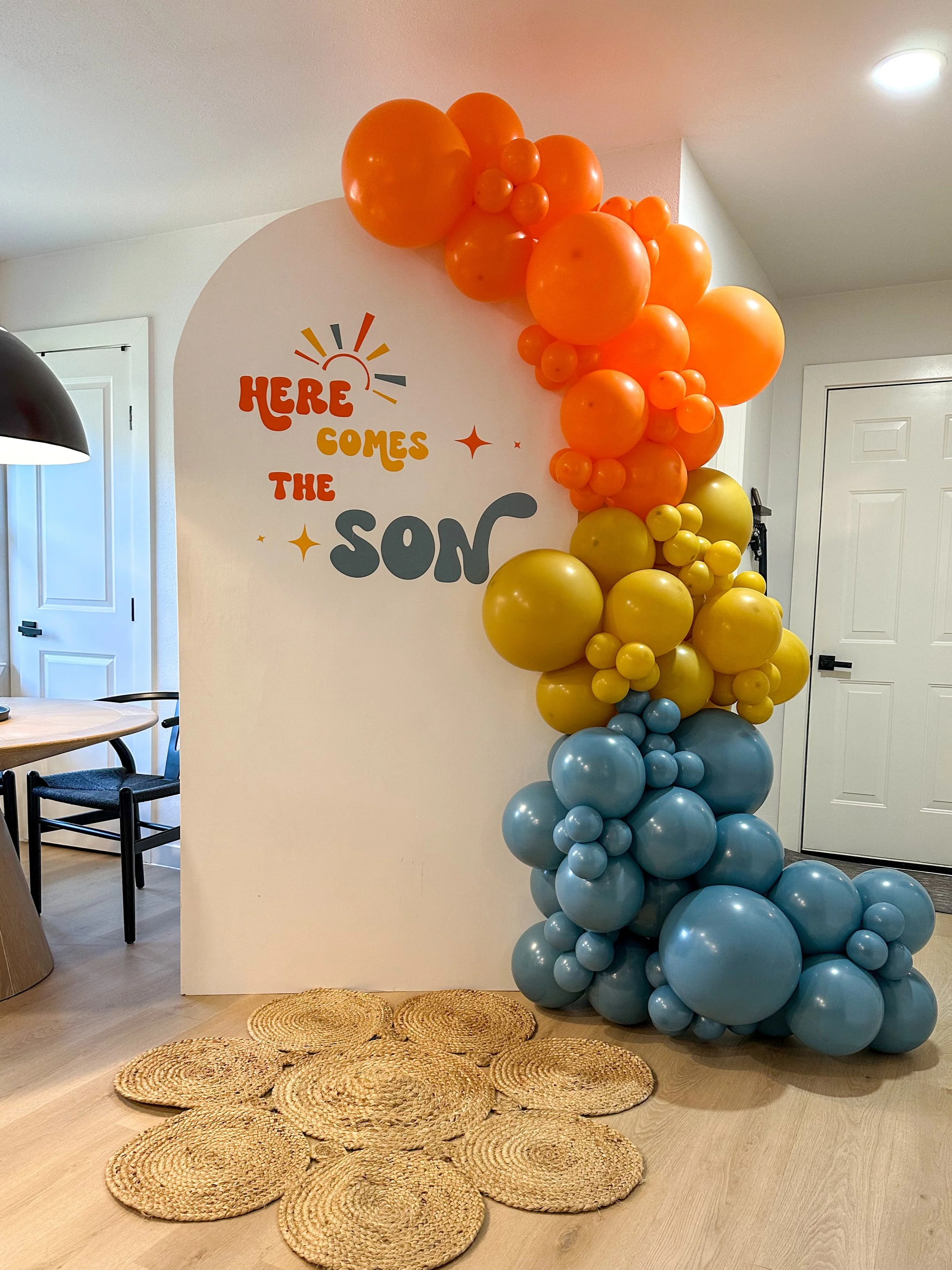 Decorative display with colorful balloons in orange, yellow, and blue stacked on a white wall with a sign that says, 'Here Comes the Son.' on a woven mat in a room with white doors and a wooden table.
