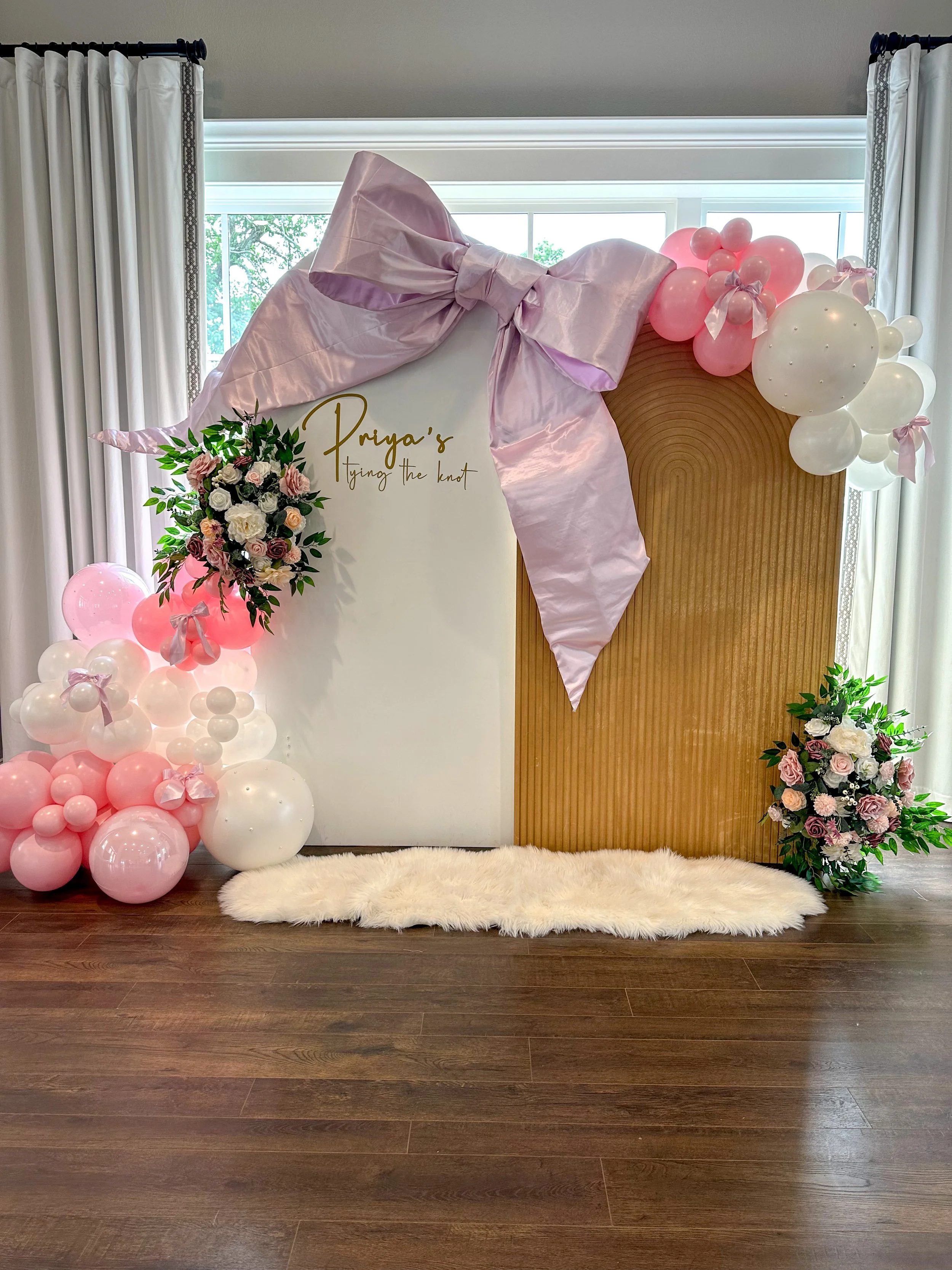 Decorative backdrop with pink and white balloons, floral arrangements, and a large pink satin bow for a celebration, with a white and wood panel background and a fluffy rug on a wooden floor.