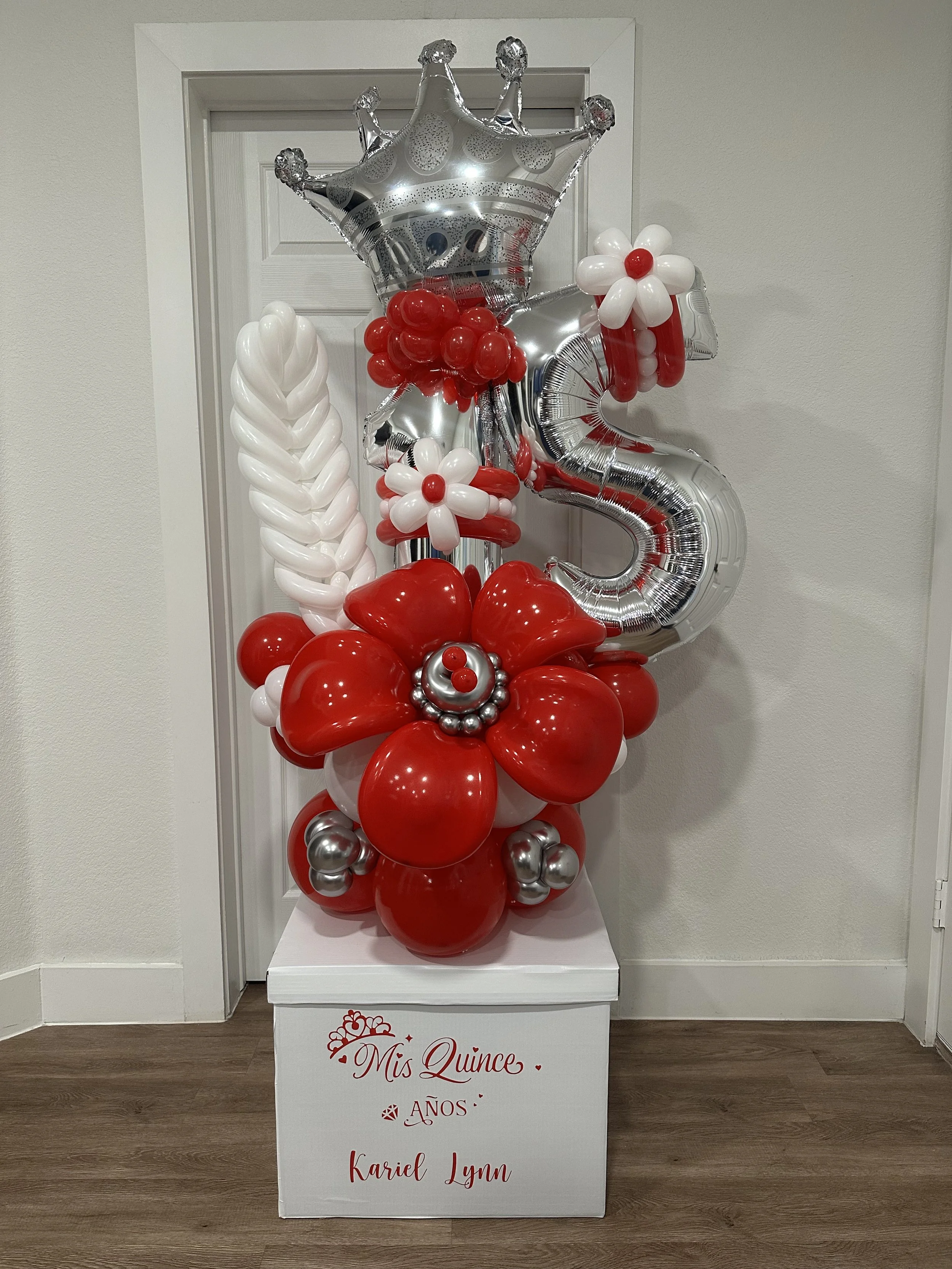 Decorative balloon arrangement for a quinceañera with a large silver number 15 balloon, including a silver crown balloon, red and white flower-shaped balloons, and additional red, white, and silver balloons, set on a white stand with red lettering th