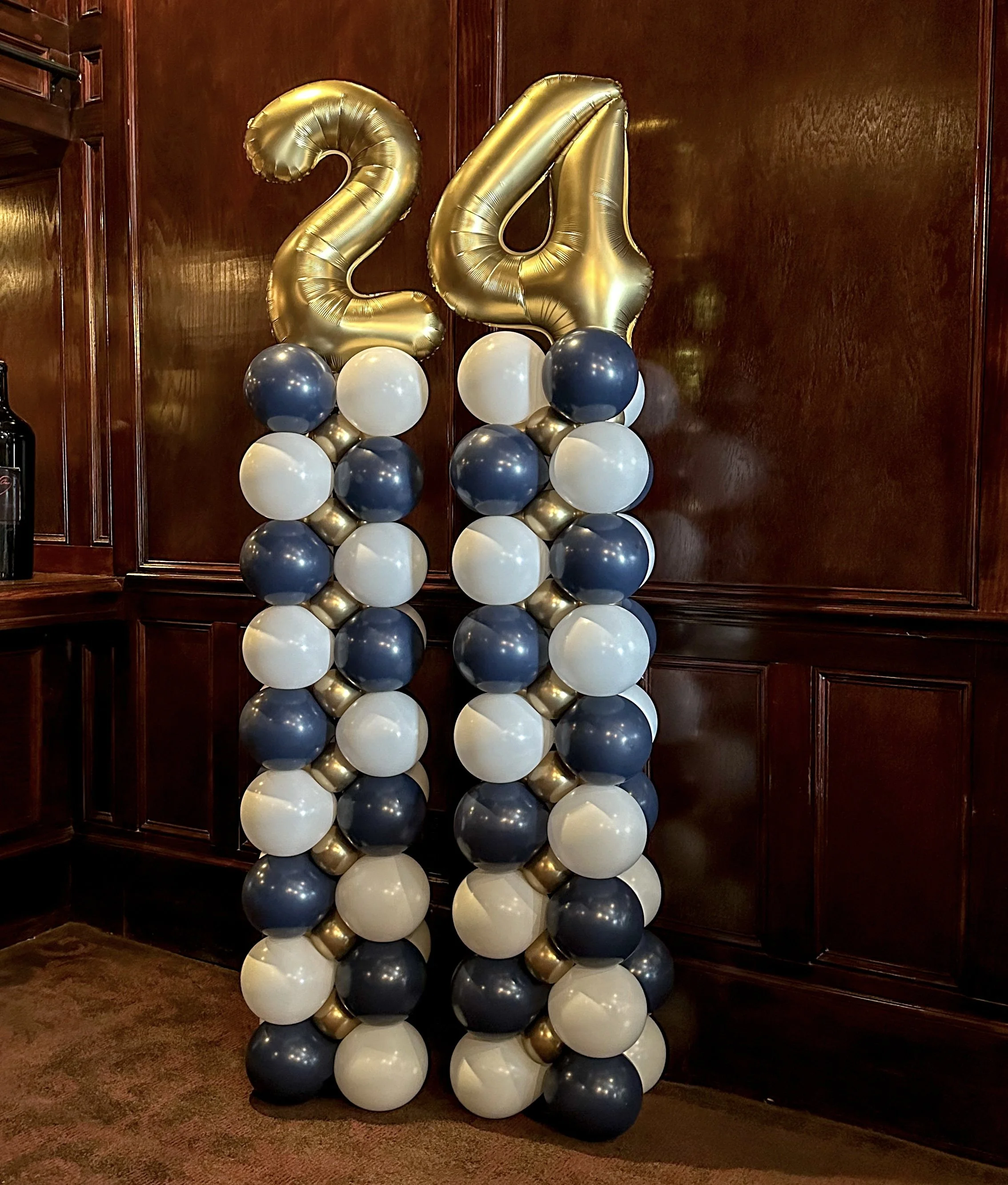 Balloon decoration with large gold number balloons '24', topped with clusters of navy blue, white, and gold balloons arranged in columns.