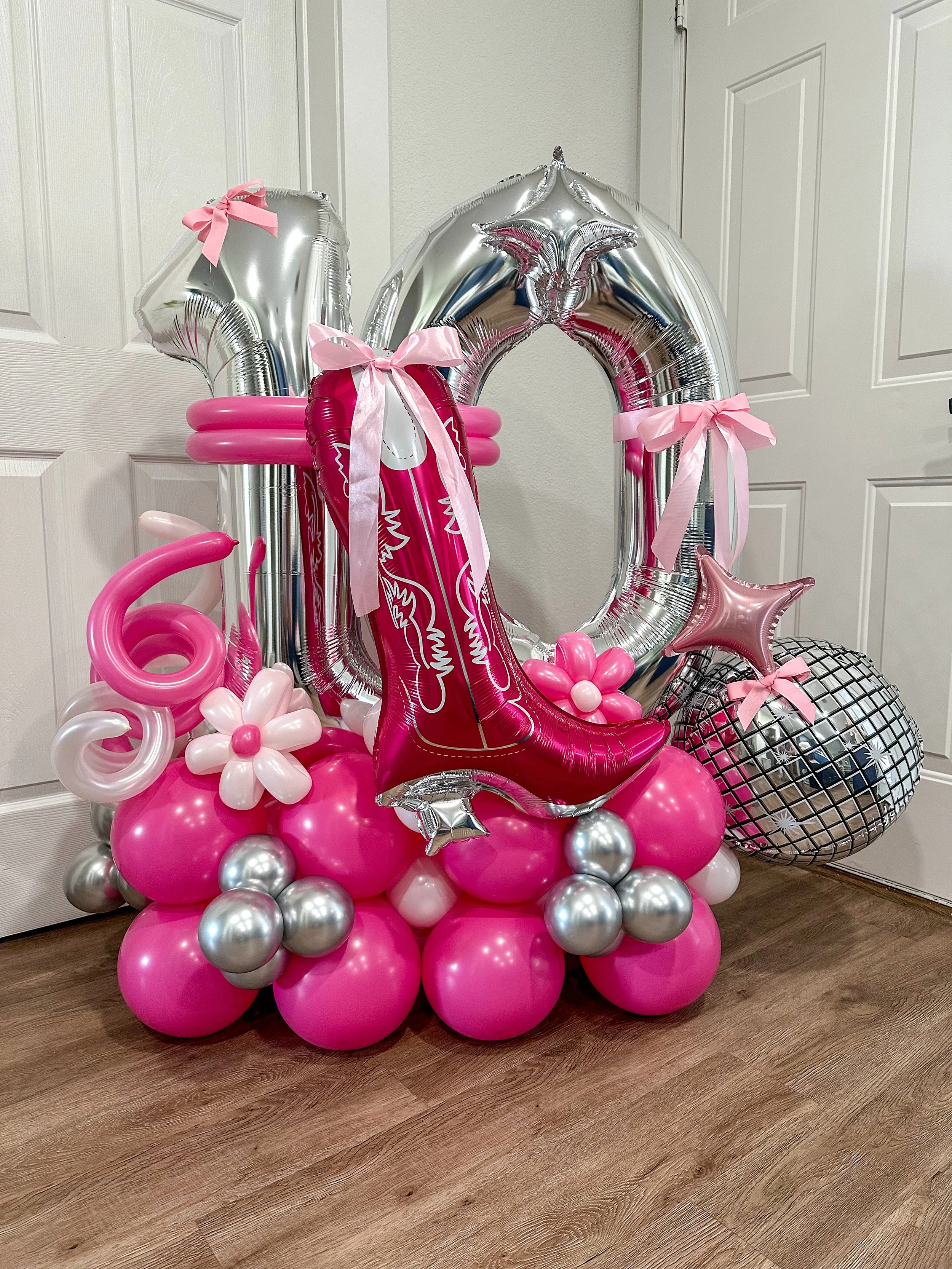 Balloon display for a 16th birthday, featuring large silver NUMERO 16 balloons, a pink cowboy boot balloon, pink and silver balloons, and a disco ball, decorated with pink ribbons and flowers.