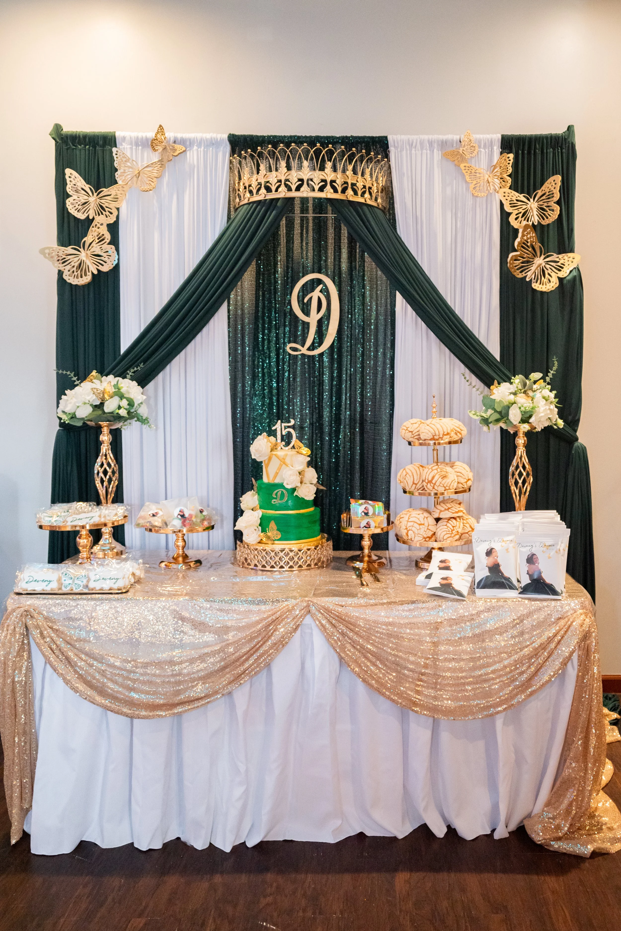 Decorated dessert table with green and gold color theme, featuring a green cake with white flowers, pastries, and printed pictures, set against a backdrop of dark green, white, and glittery black curtains with gold butterfly decorations and a gold cr
