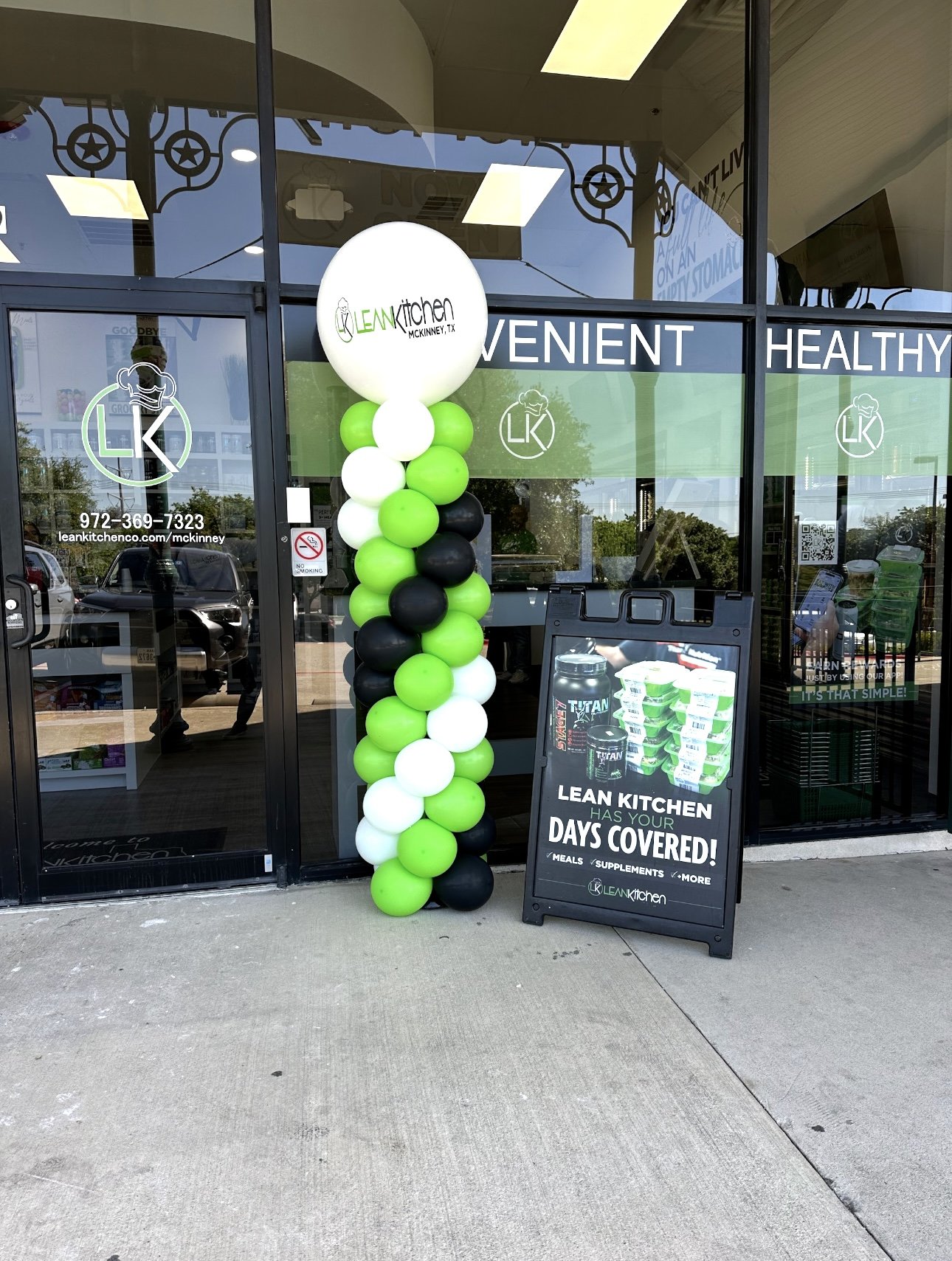 Balloon column with white, green, and black balloons outside the entrance of Leankitchen in McKinney, Texas. Signboard advertising meals, supplements, and more.