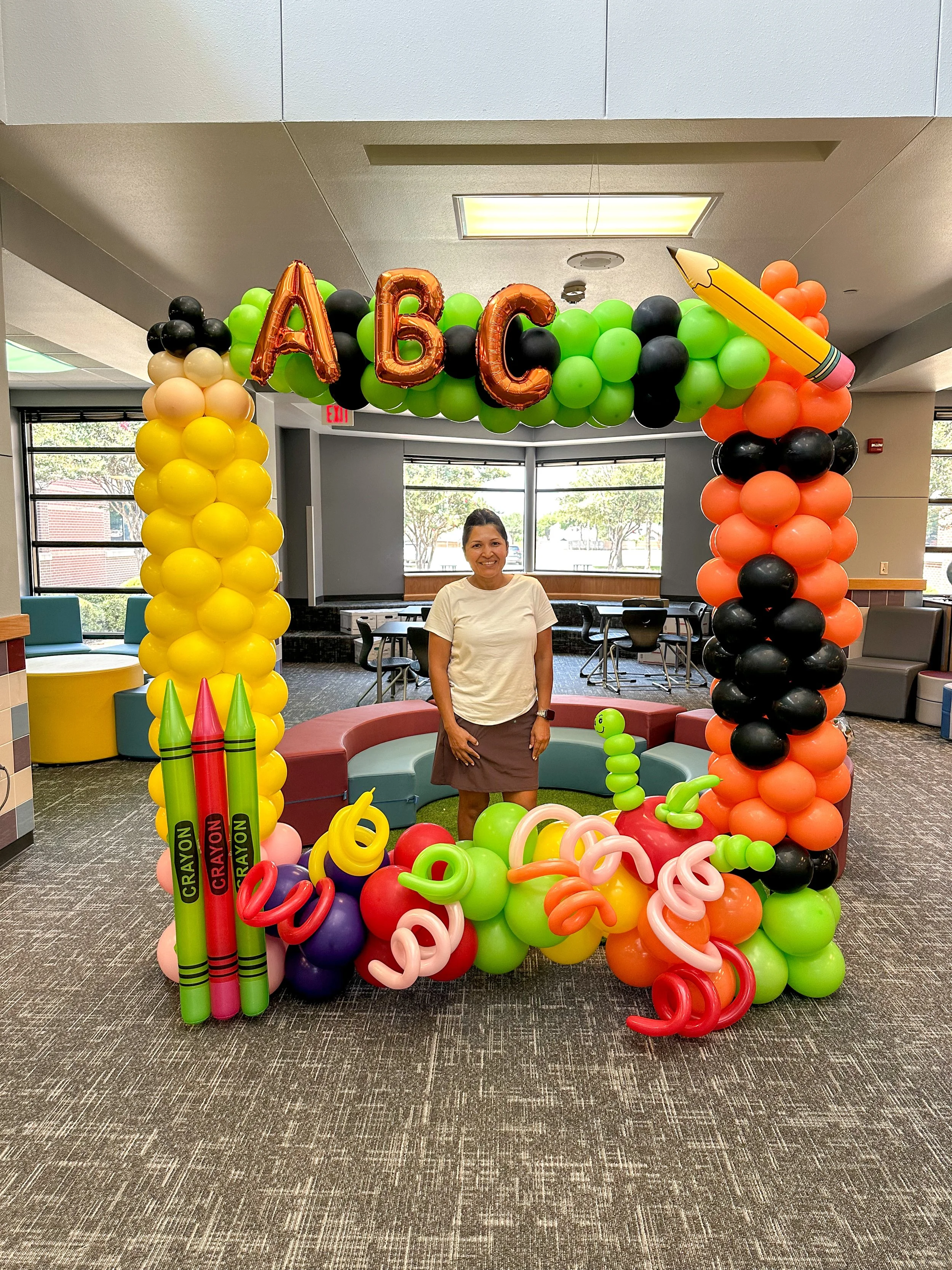 Colorful balloon arch with letters 'A', 'B', and 'C', and school supplies like crayons and a pencil, set up in an indoor space. A woman stands inside the arch.