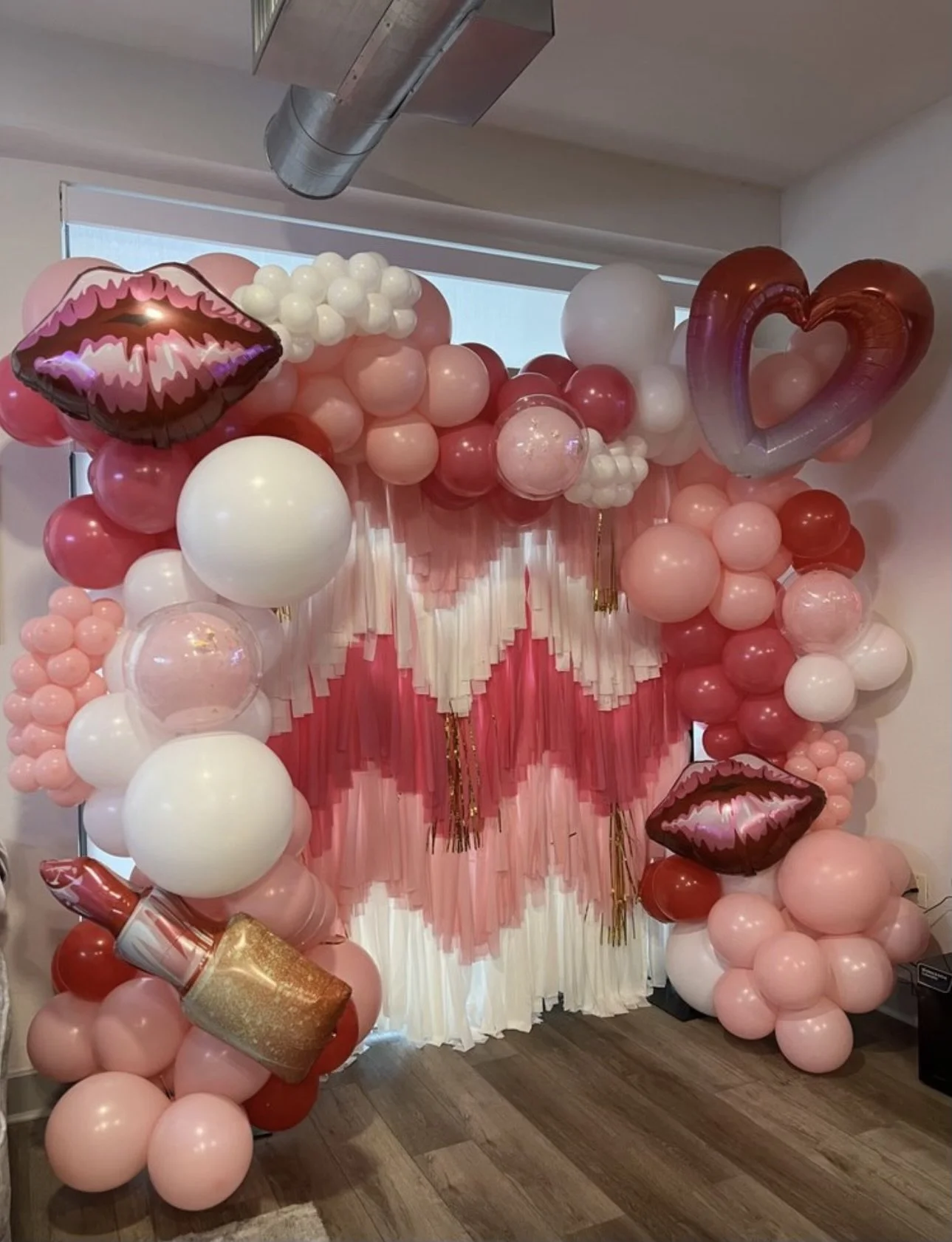 Decorative backdrop with balloons in pink, white, and red, including lips, lipstick, and heart-shaped balloons, ideal for a celebration or photo booth.