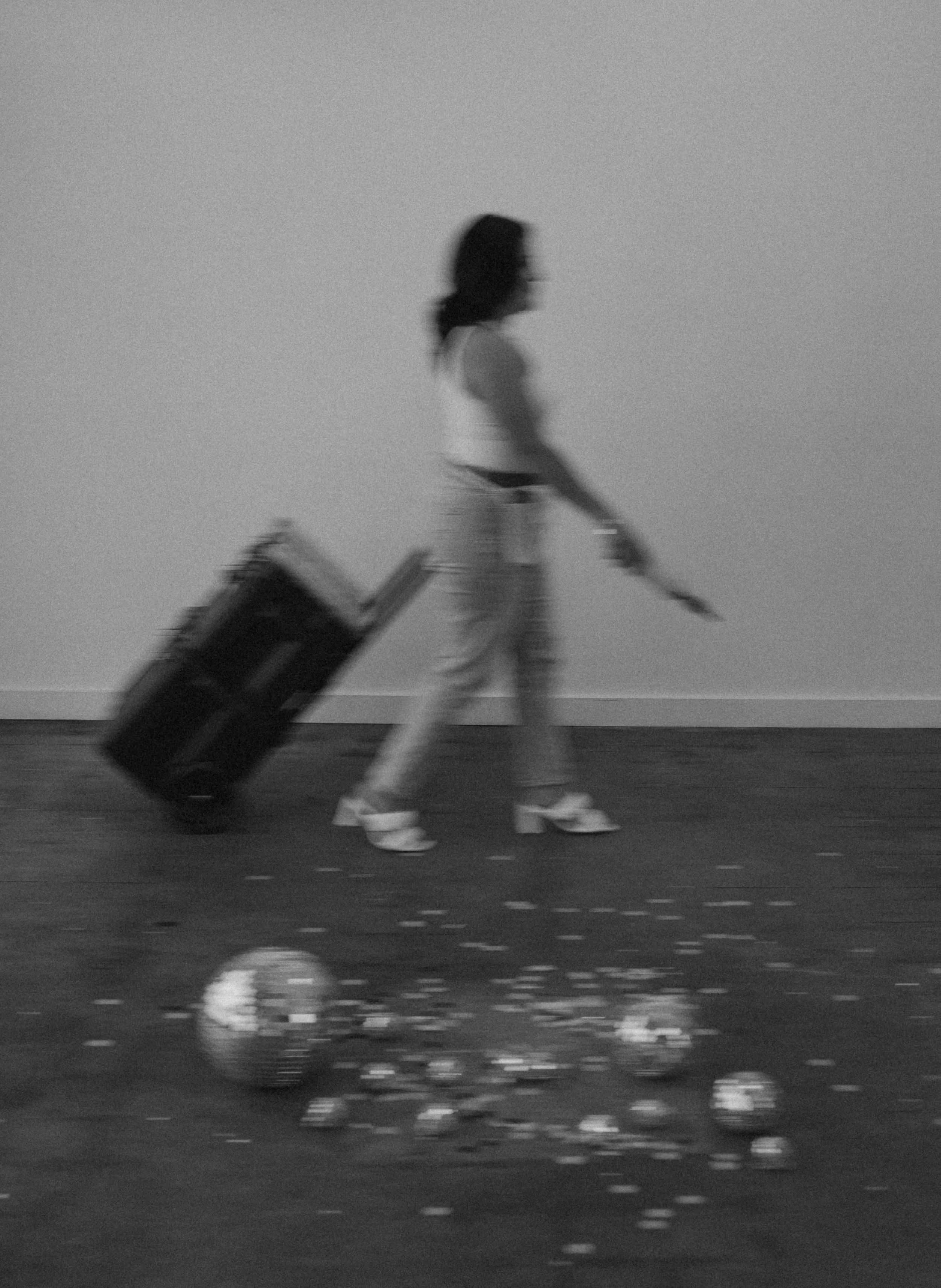 A woman walking with a rolling suitcase and a skateboard in an empty room, with broken glass and debris on the floor, black and white photograph.