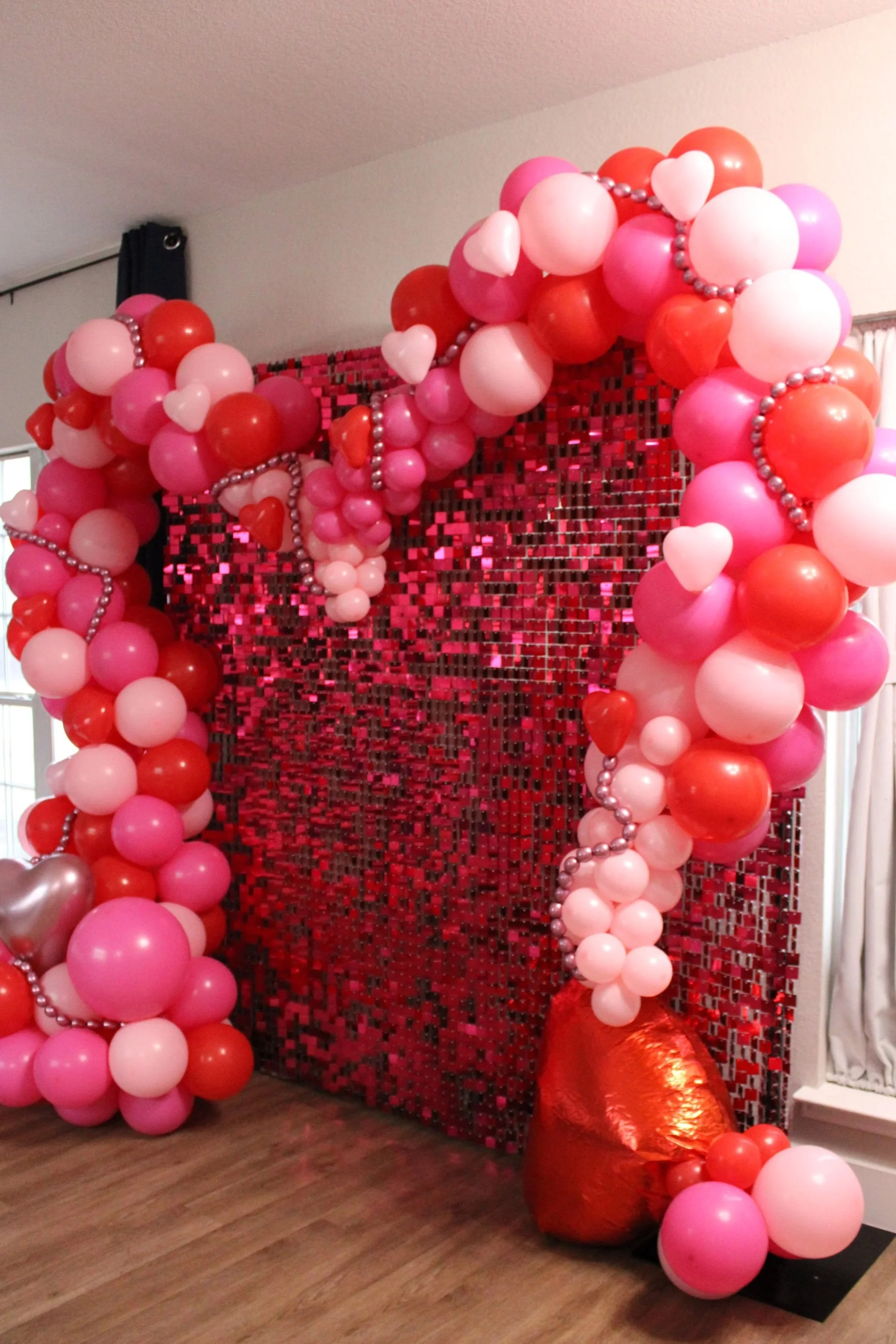 Decorative backdrop with pink, red, and white balloons arranged in an arch and cluster, with a shiny red heart balloon at the bottom, set against a sequin-covered wall for a celebration or Valentine's Day event.