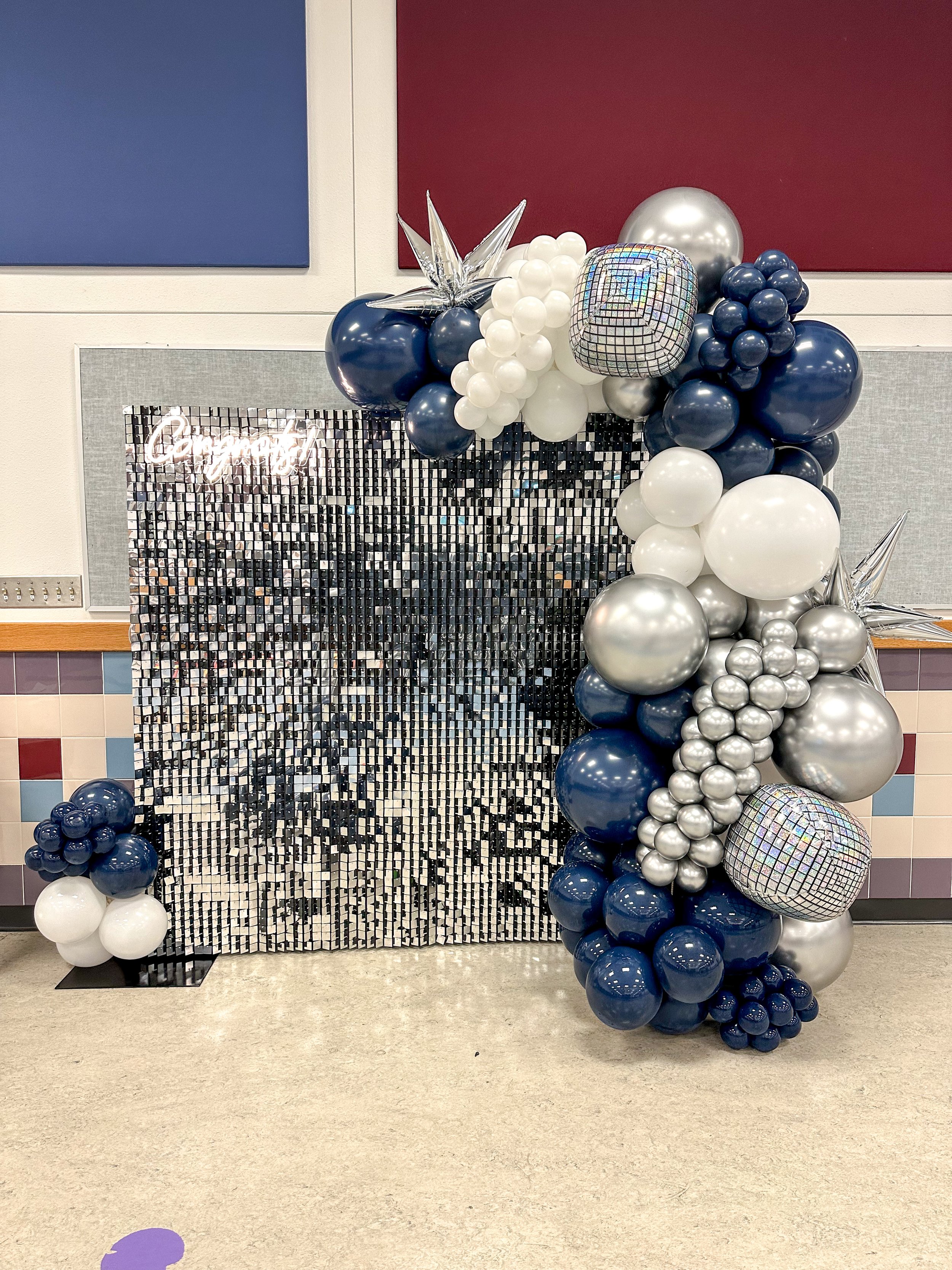 Decorative balloon arrangement with blue, white, silver, and holographic balloons around a black and silver mirrored backdrop.