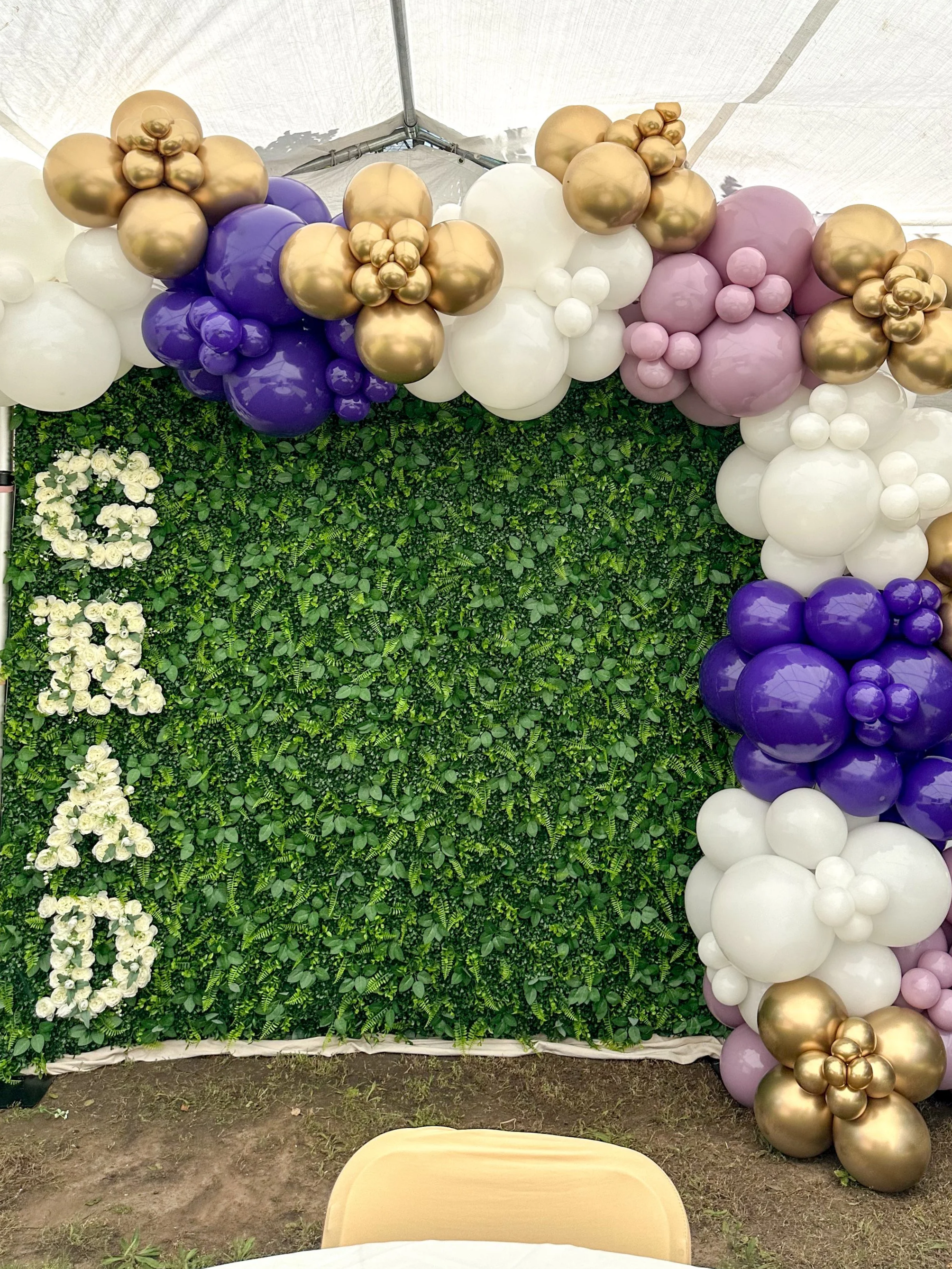 Decorative backdrop with a green leafy wall, white floral letters spelling out 'SOY', surrounded by purple, white, gold, and pink balloons forming an arch.
