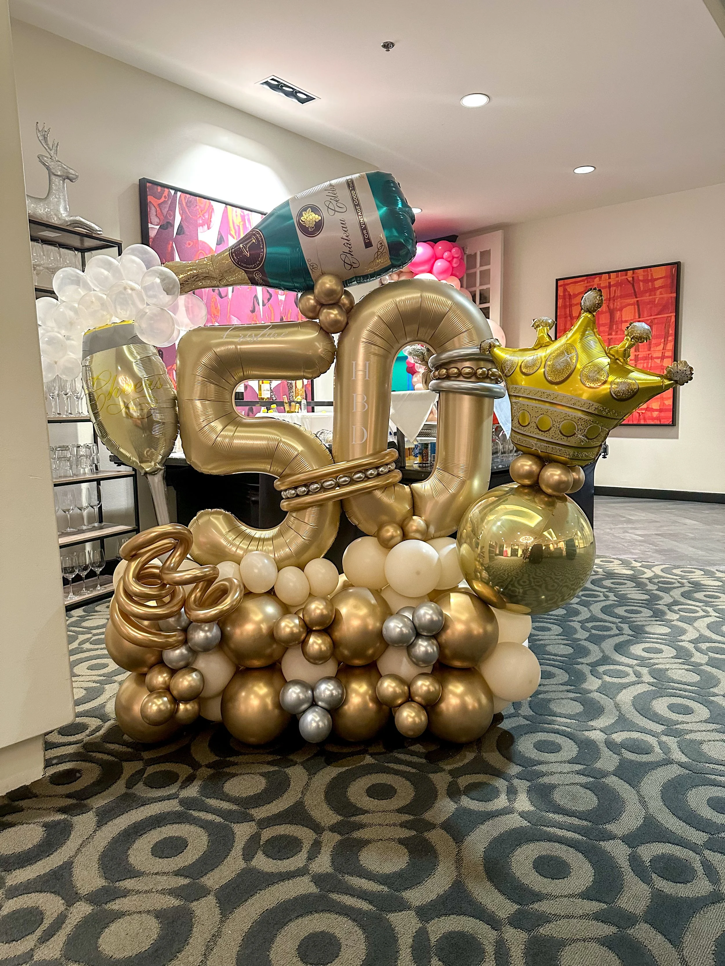 Balloon decoration for a 50th birthday celebration featuring large gold and white balloons, a champagne bottle balloon, a gold crown balloon, and a cluster of smaller balloons at the base.