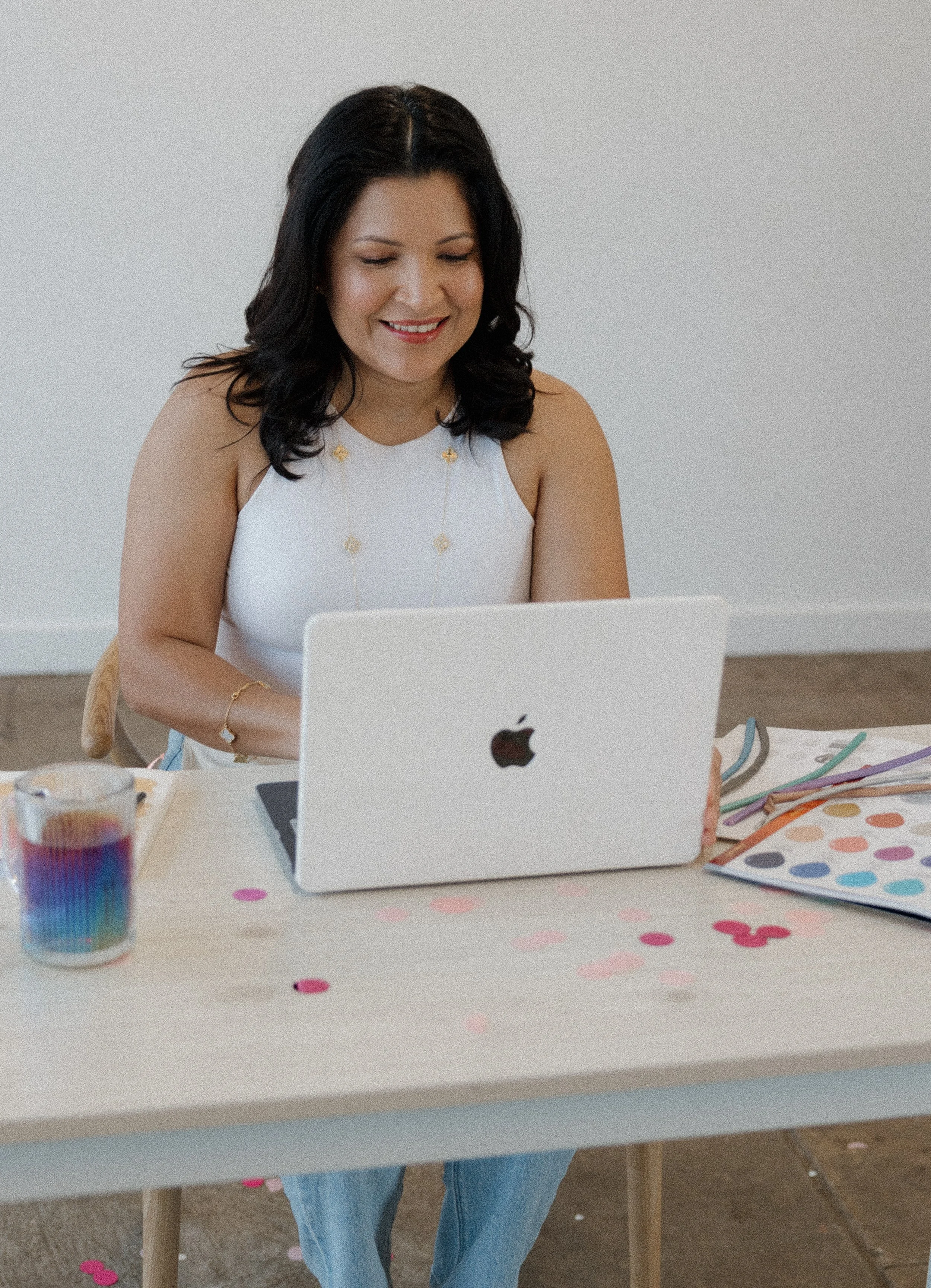 A woman with black hair and light skin smiling while sitting at a table with a silver MacBook laptop, colorful paper confetti, and a rainbow-colored drink in a glass.