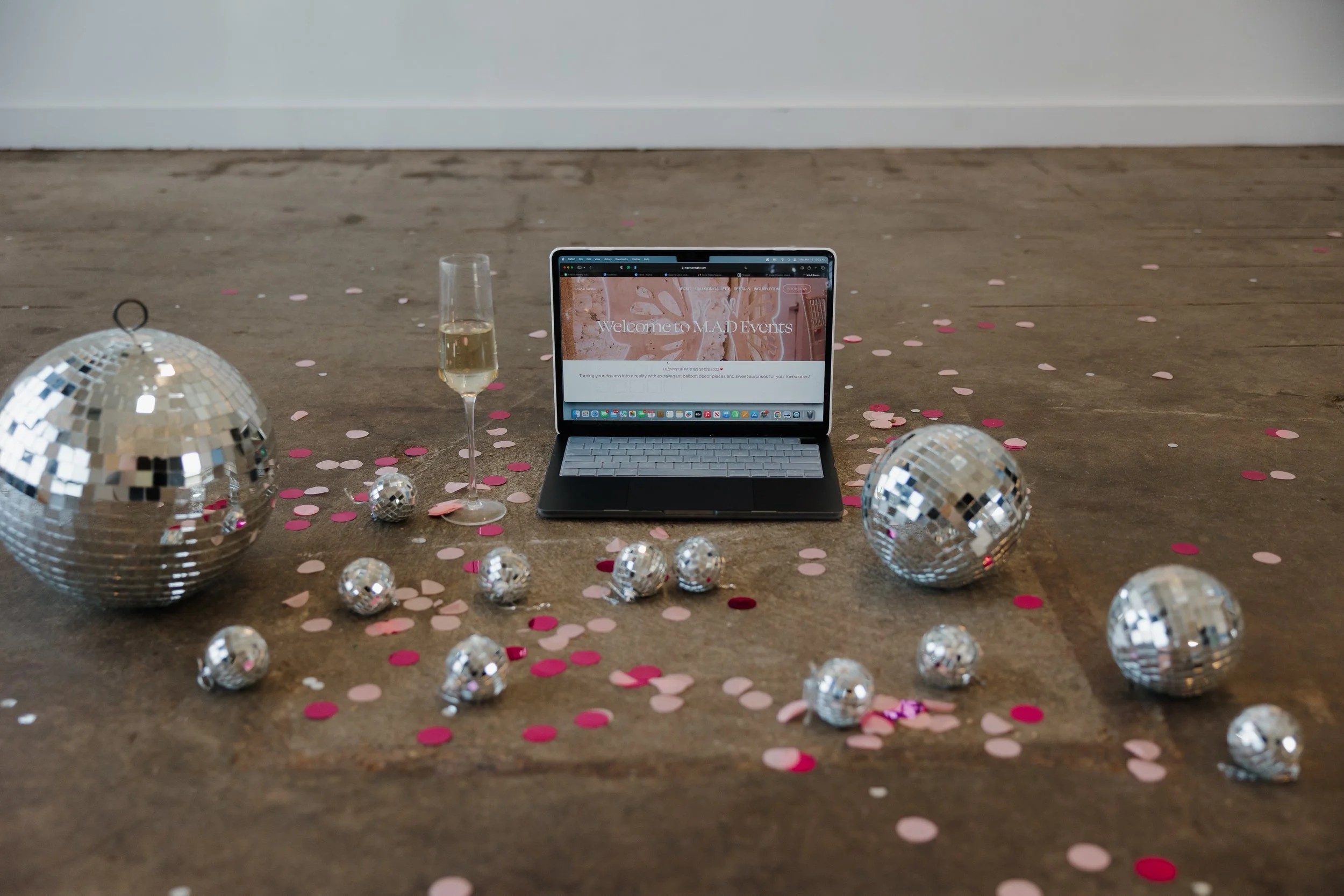Decorative celebration setup with a laptop displaying 'Welcome to M.A.D Events', two disco balls, a glass of champagne, and pink and white confetti scattered on a wooden floor.