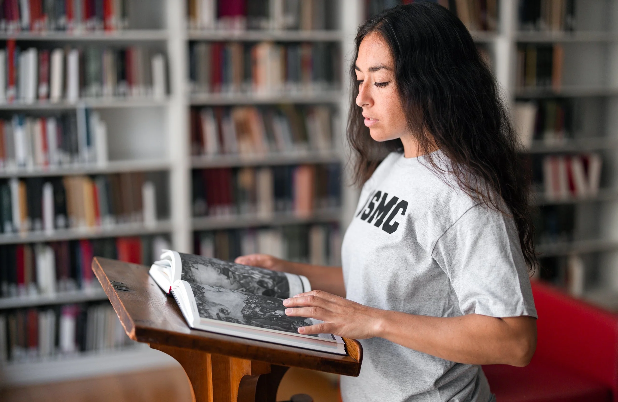 A woman in a white USMC shirt reading a book that sits on a wooden podium