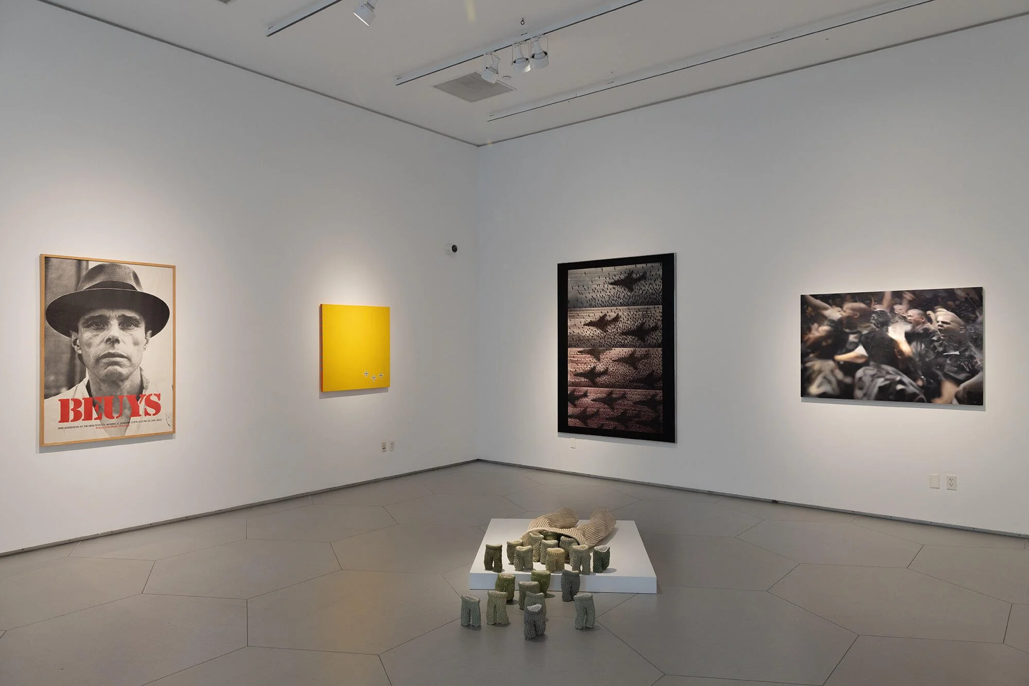 An art gallery with white walls, displaying four framed artworks: a black-and-white photo of a man in a hat with red text, a yellow abstract piece, an artwork with birds flying in formation over a body of water, and a picture of people and a dog at a crowded event. In the foreground, there is a white table with various small, gray, textured objects. The room has white ceilings with track lighting.