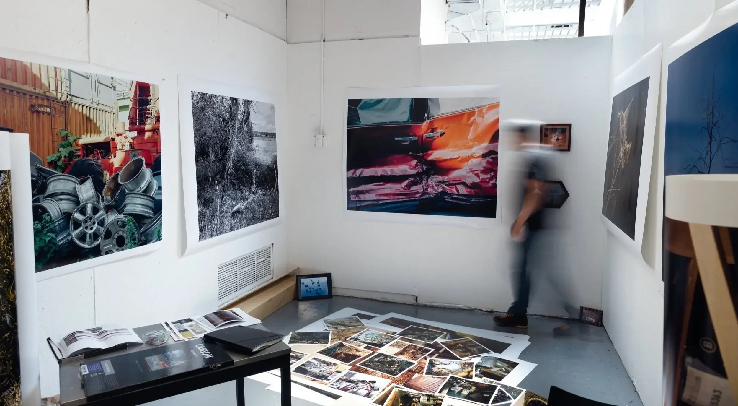 An art gallery with various large photographs on white walls, some on the floor, and a small table with photo books and stacks of photographs.