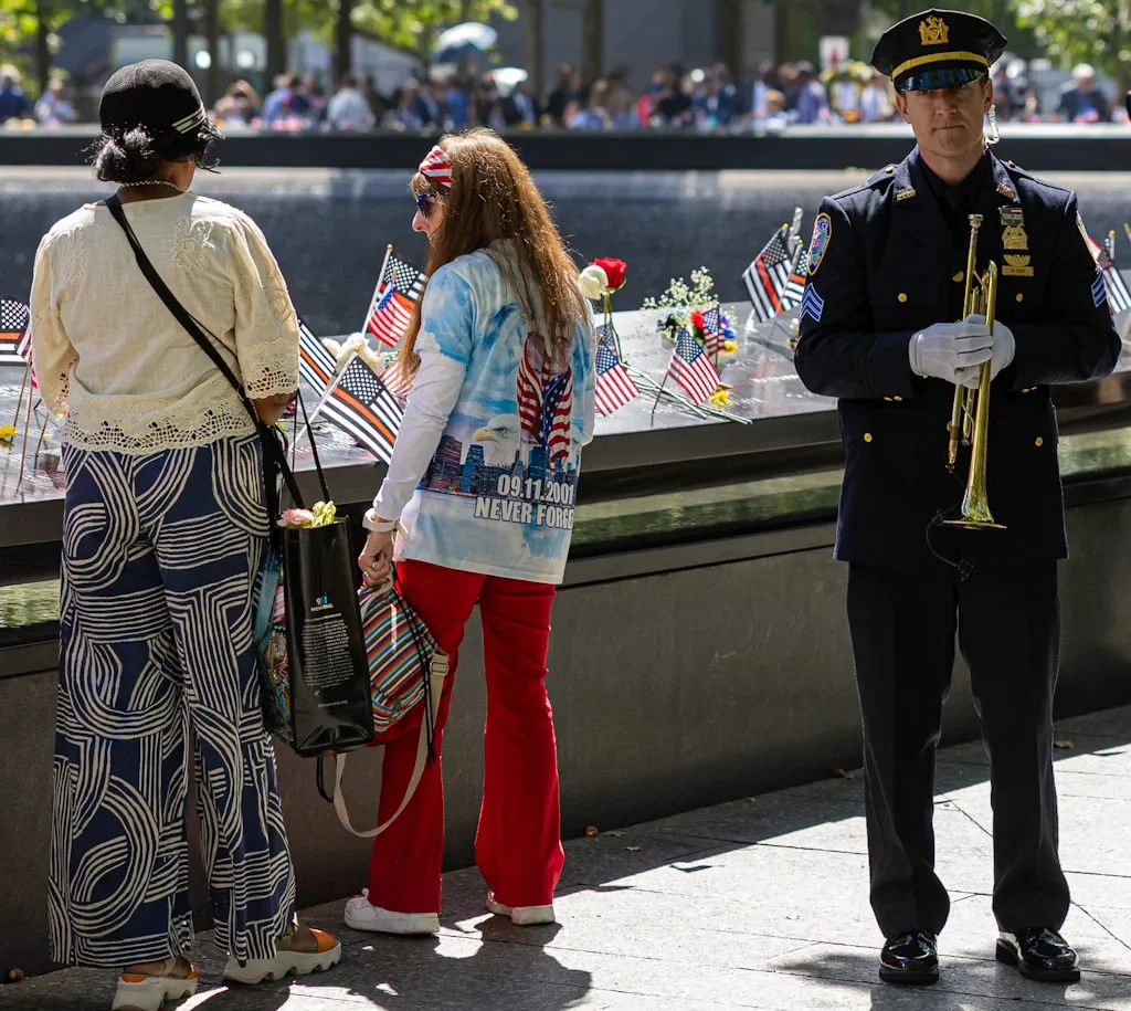 Police bugler in dress uniform standing beside visitors at the 9/11 Memorial pools on the 25th anniversary