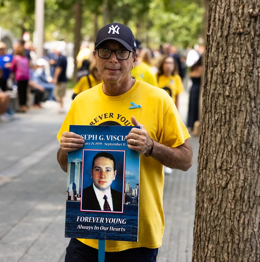 Man holding a Forever Young tribute poster for a 9/11 victim at the 25th anniversary ceremony in New York City