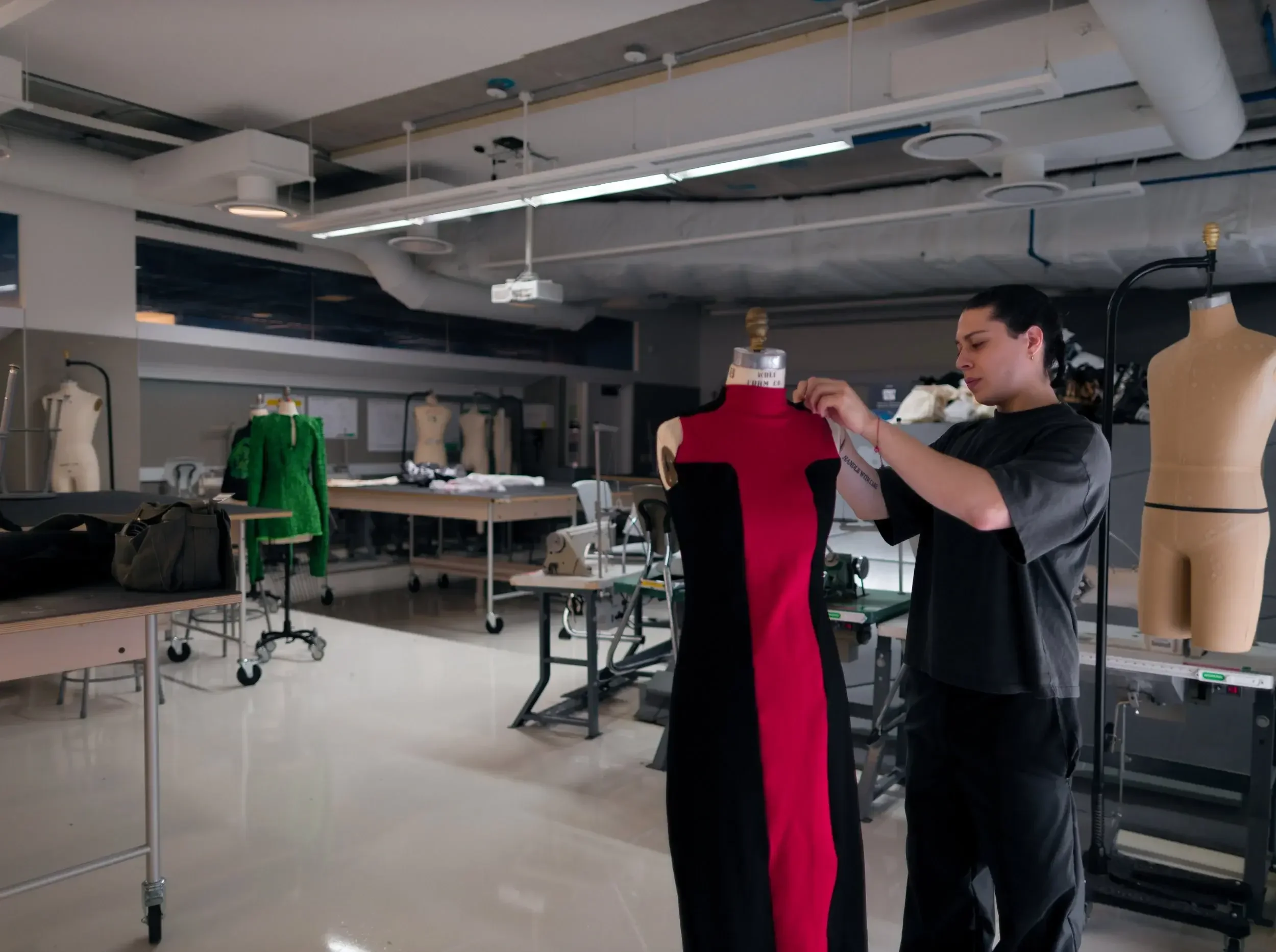 A woman adjusting a red and black dress on a dress form in a fashion design studio with mannequins, fabric, and sewing equipment.