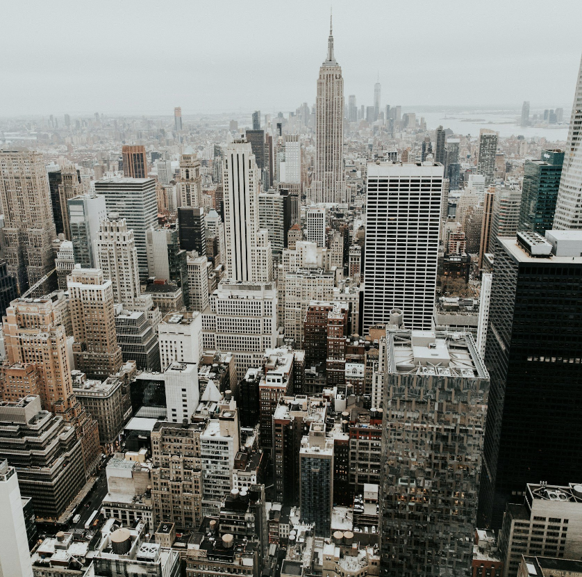 A cityscape of New York City showing numerous tall buildings and skyscrapers, with the Empire State Building prominently in the center and the Hudson River visible in the distance.