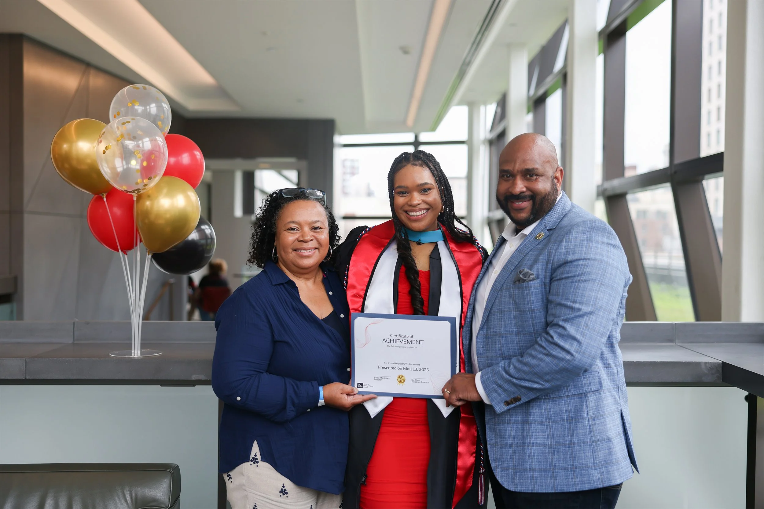 A young woman in a graduation gown holding a certificate, smiling with her parents who are standing beside her. There are balloons in the background.