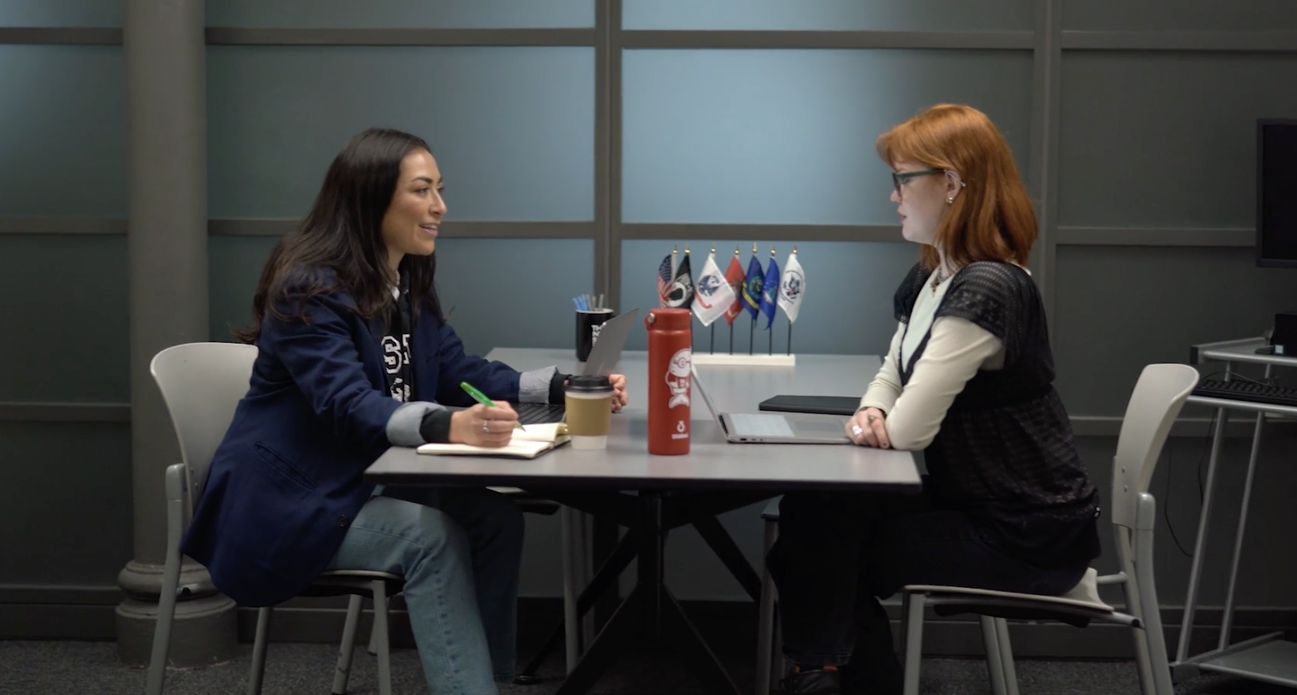 Two women sitting at a table in a professional office setting having a conversation.