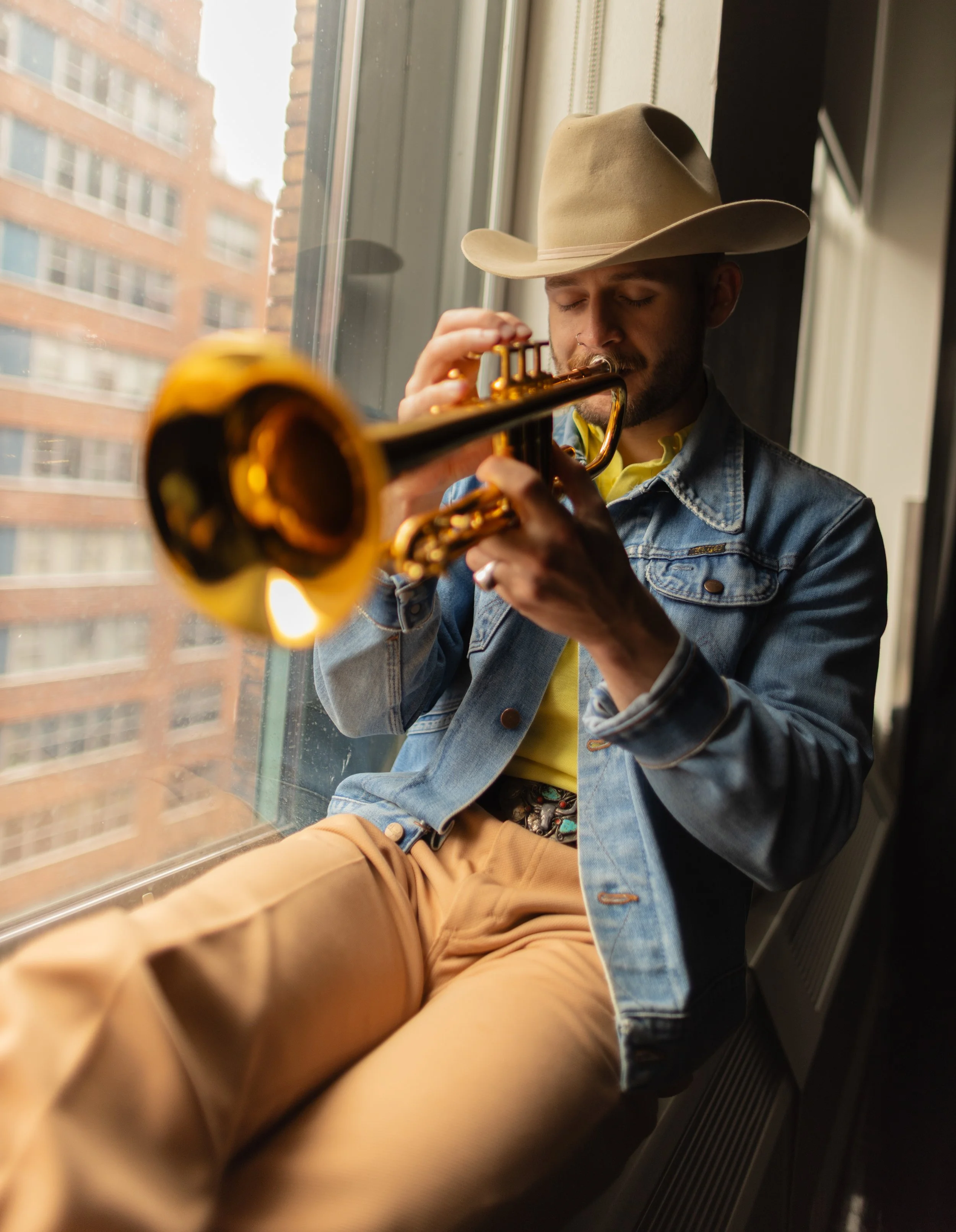 Austin Simmons playing trumpet by a window overlooking New York City