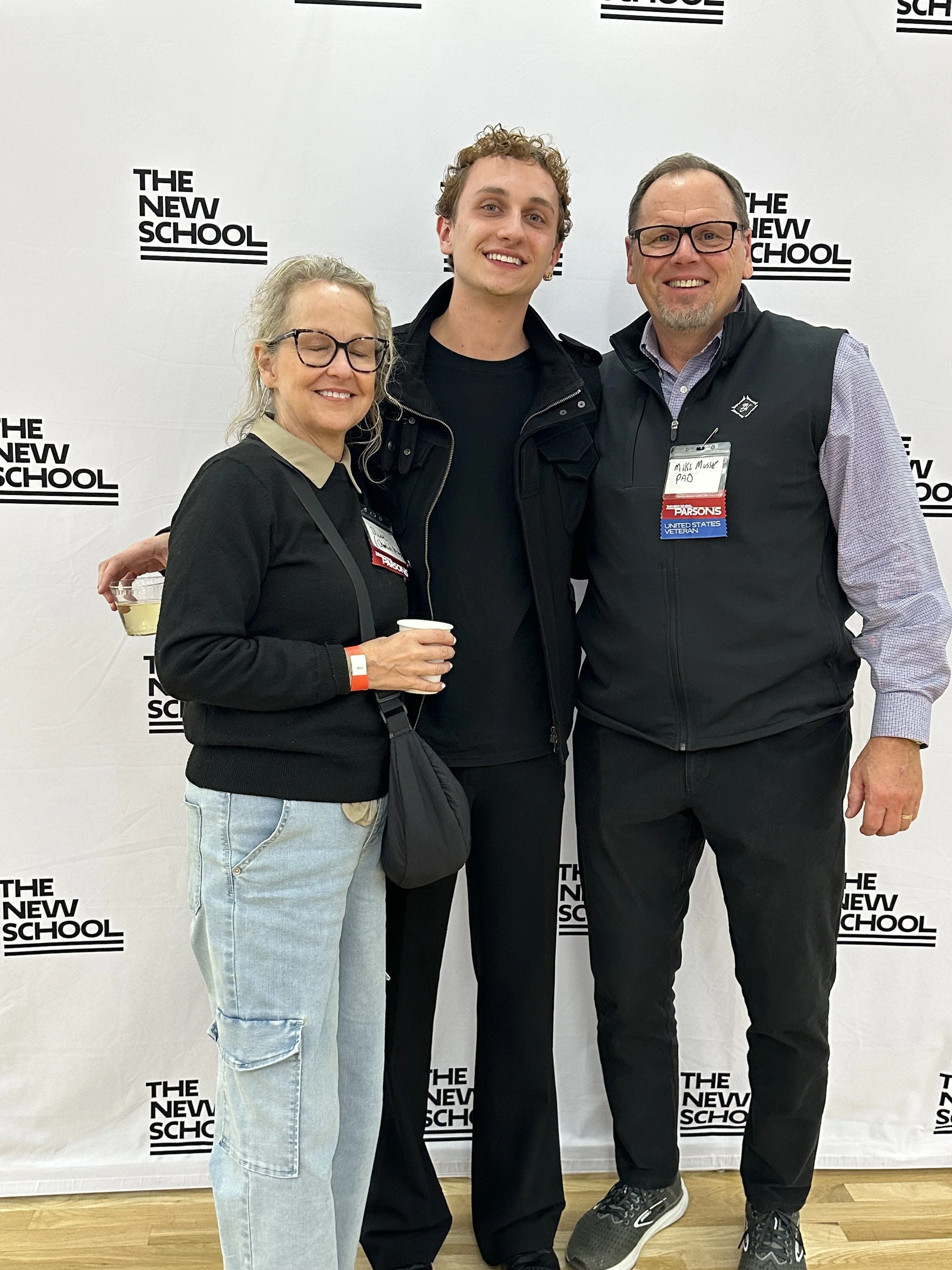 Three people smiling and posing for a photo in front of a backdrop that reads 'The New School.' The woman on the left has blond hair, glasses, and is holding a glass of white wine. The young man in the center has curly hair and is wearing a black jacket. The man on the right has short gray hair, glasses, and is wearing a vest over a button-up shirt.