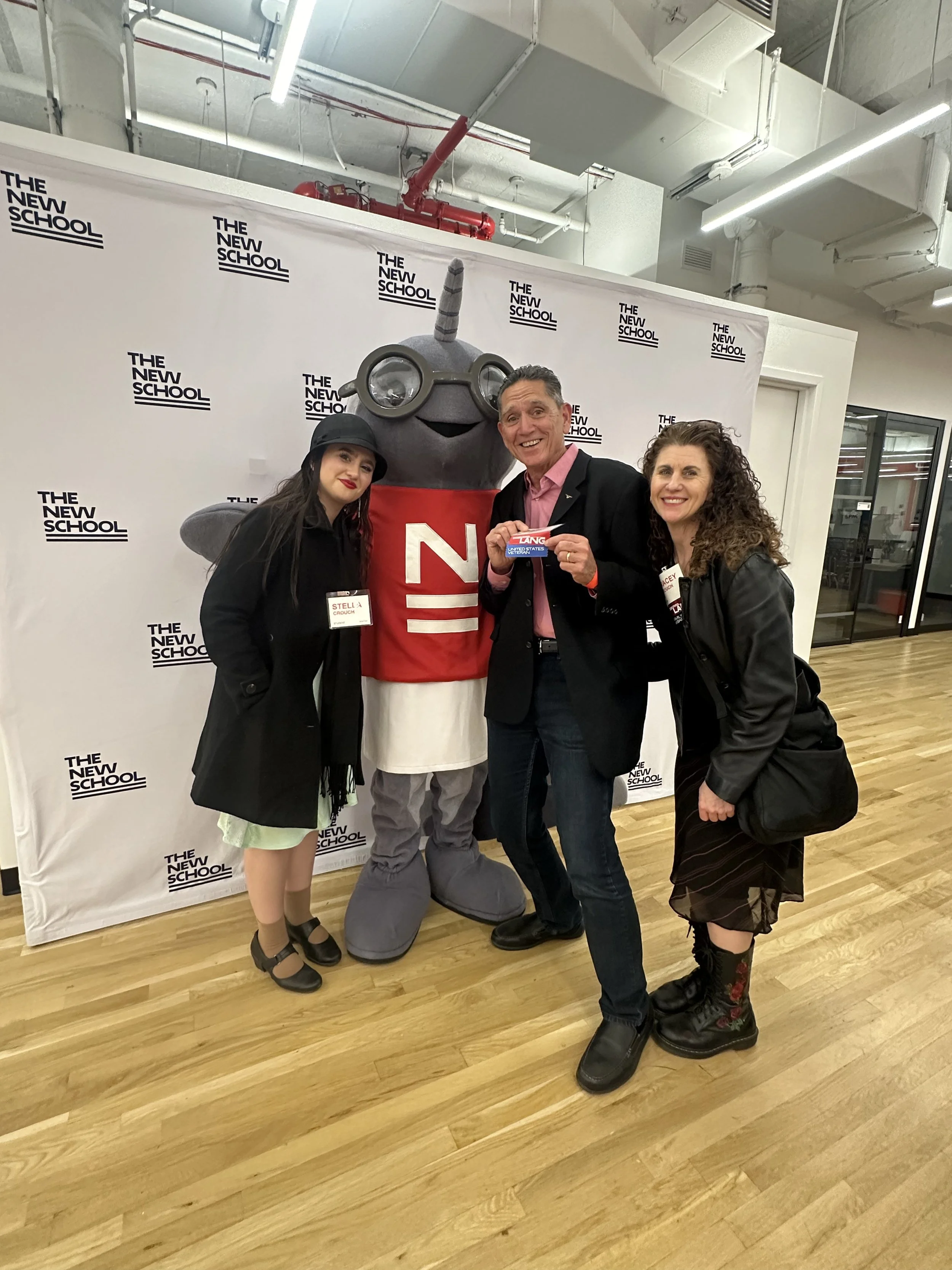 Four people standing with a mascot in front of a backdrop that reads 'The New School'. The mascot is a gray unicorn wearing goggles and a red shirt with a white 'N'. One person is holding a sign that says 'VANG'. All are smiling and dressed in casual or semi-formal attire, indoors with a wooden floor and industrial ceiling.