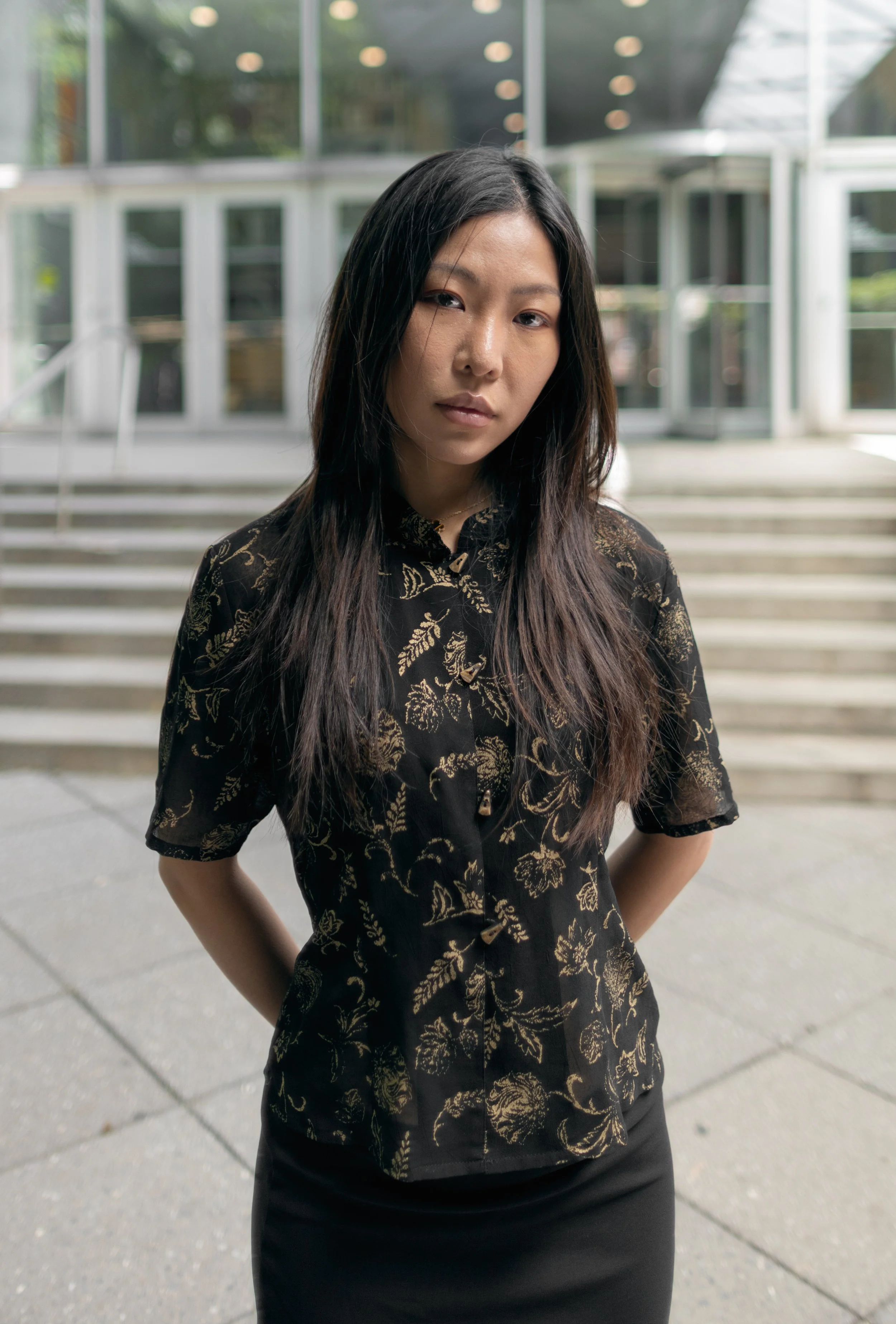 Young woman with long dark hair wearing a black floral patterned short-sleeve shirt and a black skirt, standing outdoors on steps in front of a glass building.