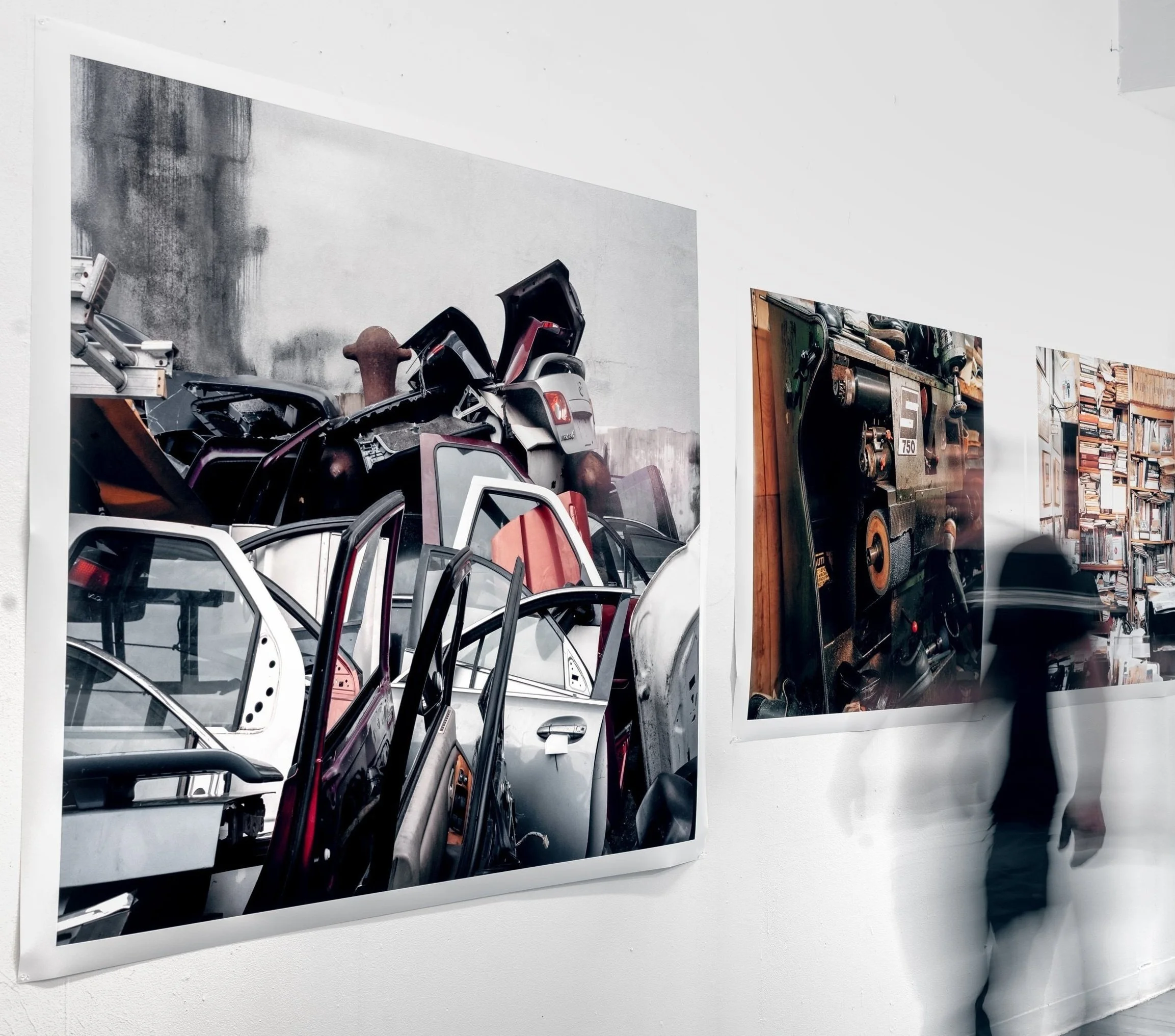 Photograph of various vintage cars and car parts displayed on white walls in a museum or gallery setting.