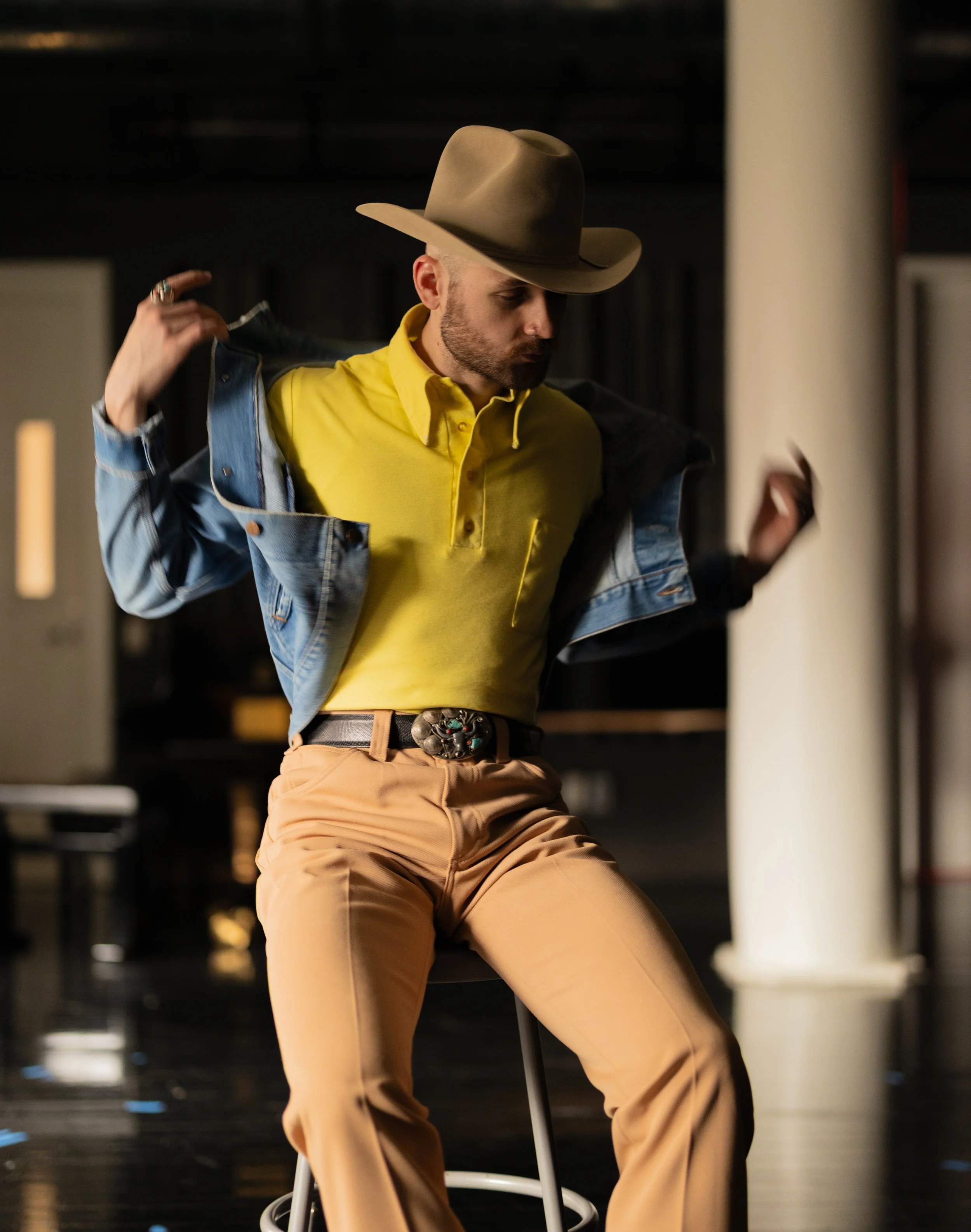 Austin Simmons posing on a stool in a yellow shirt, denim jacket and cowboy hat
