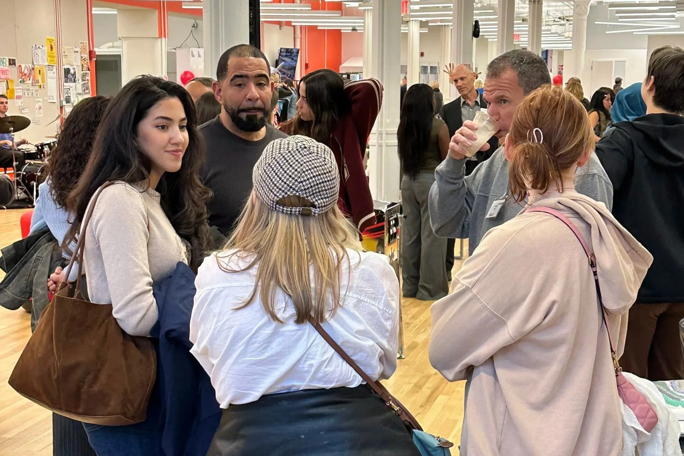A group of diverse people gathered in a busy indoor space, some talking and one man drinking from a clear cup, with a drummer playing in the background.