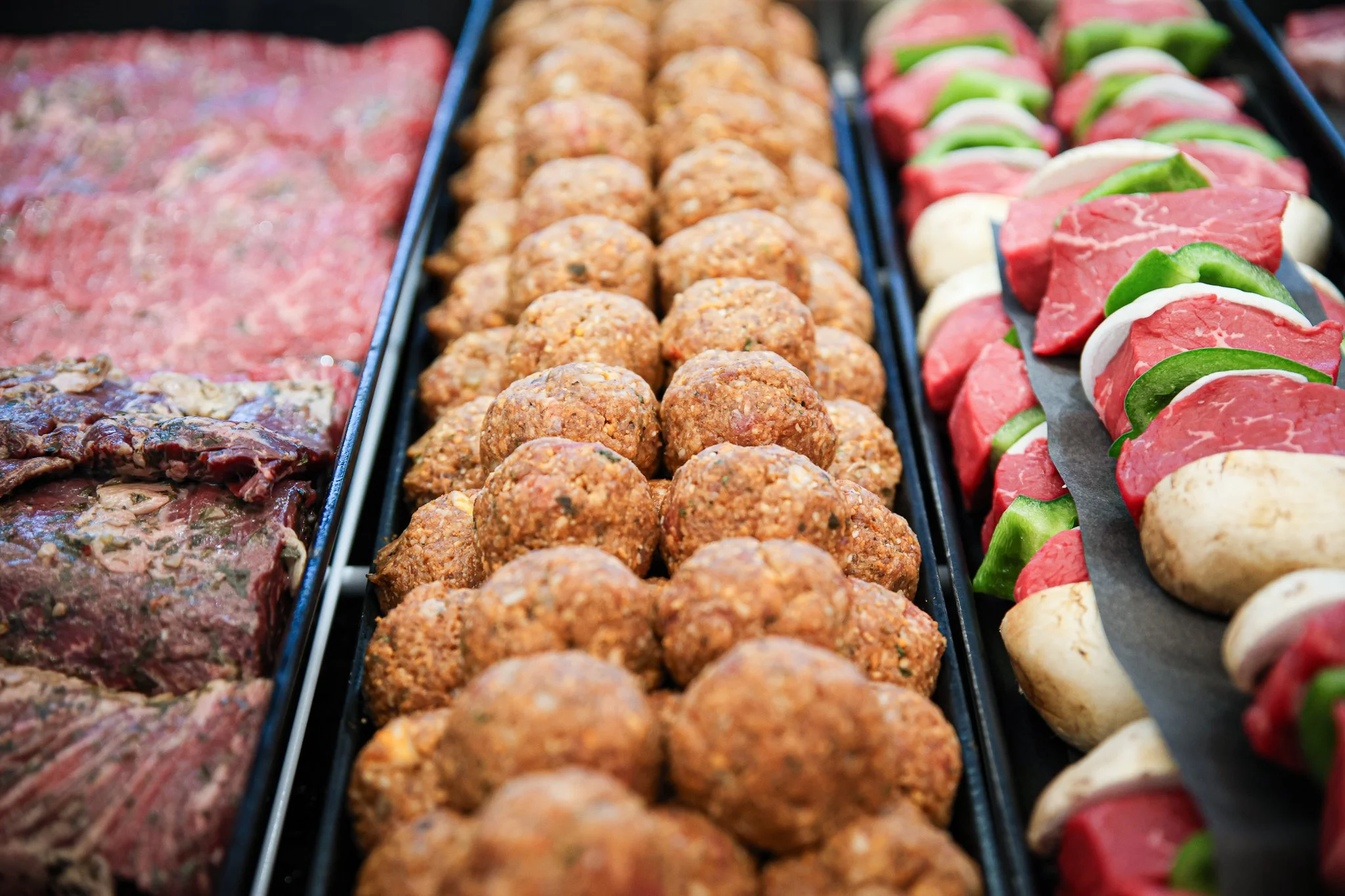 Display of raw meat, cooked meatballs, and skewered caprese salad at a deli or cookout.