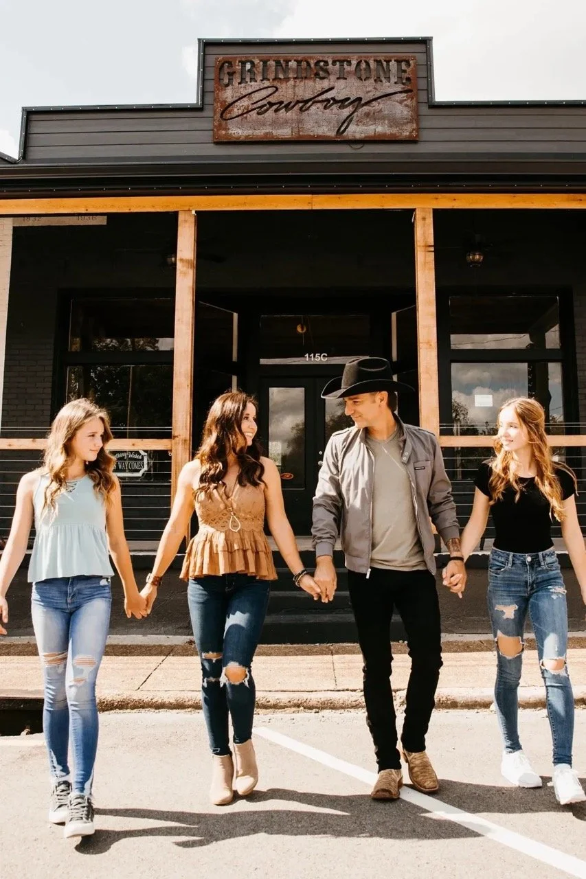 Four young women and a young man holding hands and walking in front of a building with a sign that reads 'Grindstone Cowboy.' The young man is wearing a black cowboy hat, gray jacket, and black pants, while the women are dressed in casual outfits with ripped jeans.