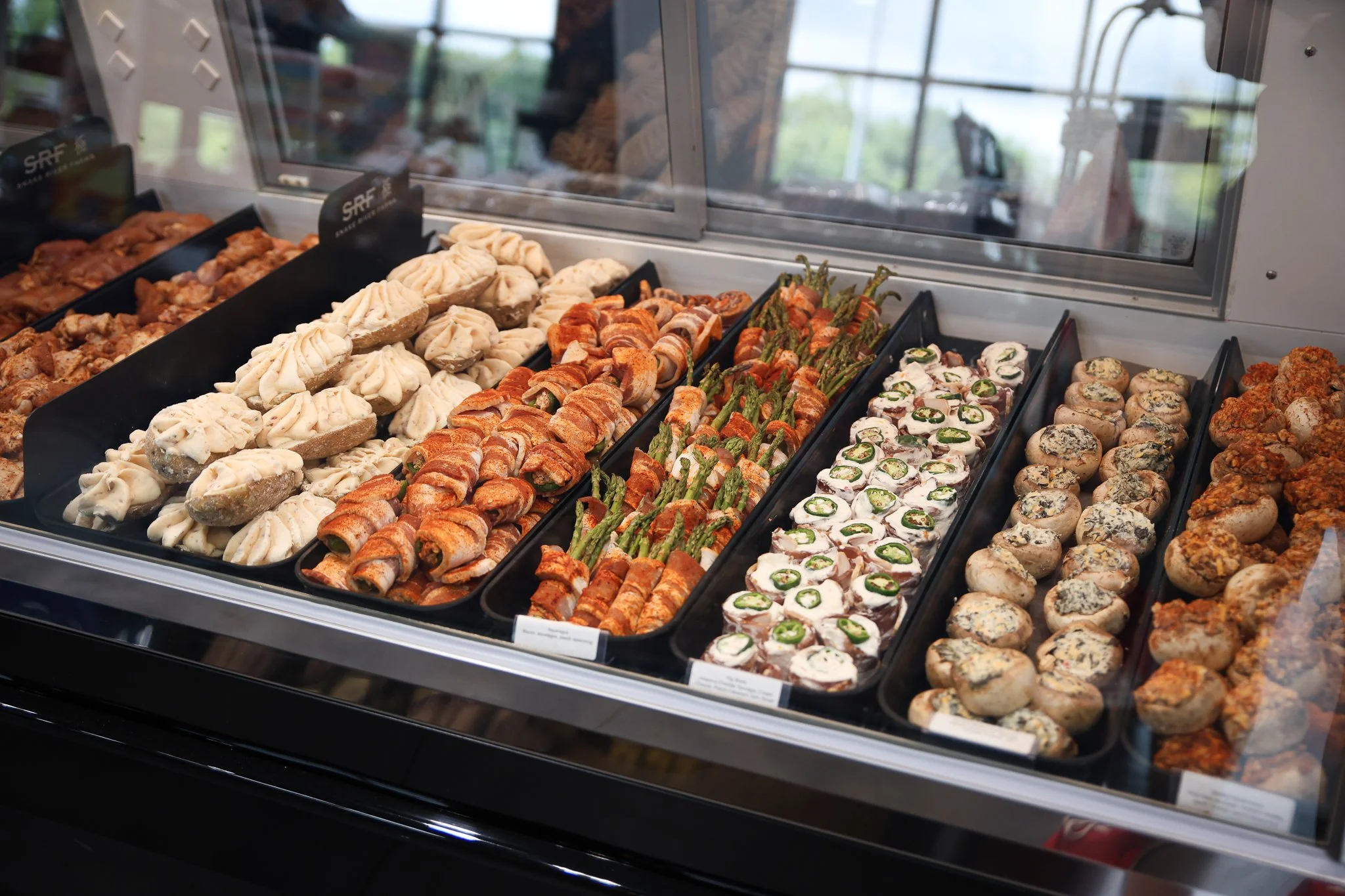 Assorted appetizers and finger foods displayed in a refrigerated case, including stuffed mushrooms, and vegetable pinwheels.
