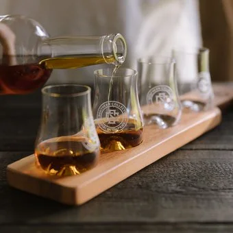 Glass tasting flight with four small glasses on a wooden tray, with one being poured from a bottle.